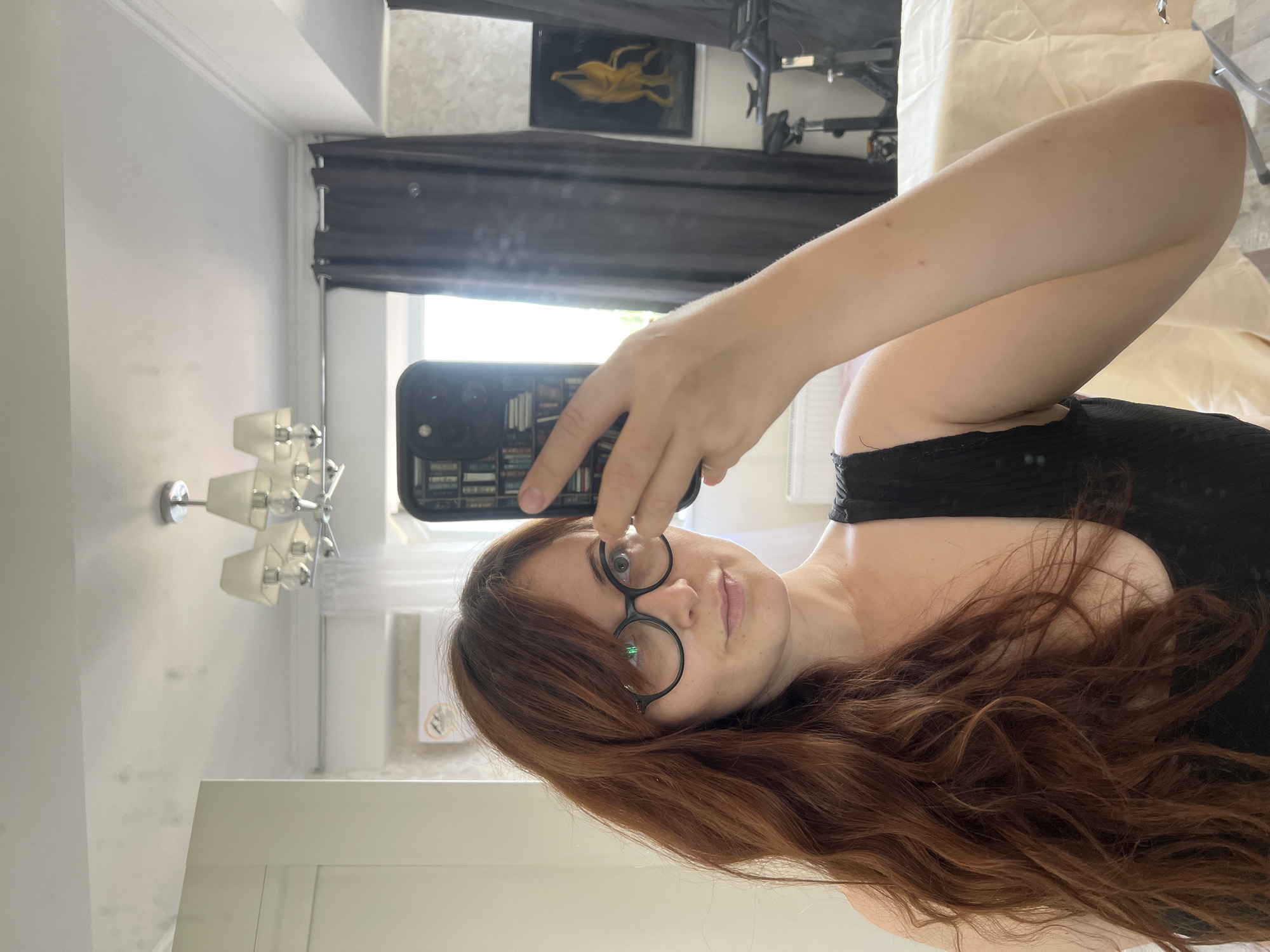 A woman with long, reddish-brown hair and round, black-rimmed glasses is taking a selfie in a mirror. She is wearing a black, ribbed tank top. Her arm is raised, holding a black smartphone with a case depicting a stack of books. The background appears to be an indoor setting, possibly a bedroom or living area, with a chandelier visible on the ceiling, dark curtains, and what looks like a painting on the wall. A bicycle ergometer is also present in the background. The image quality is moderately clear, and the lighting suggests it was taken during the daytime. The scene is devoid of any specific identifying markers that would place it in Chișinău, Moldova.