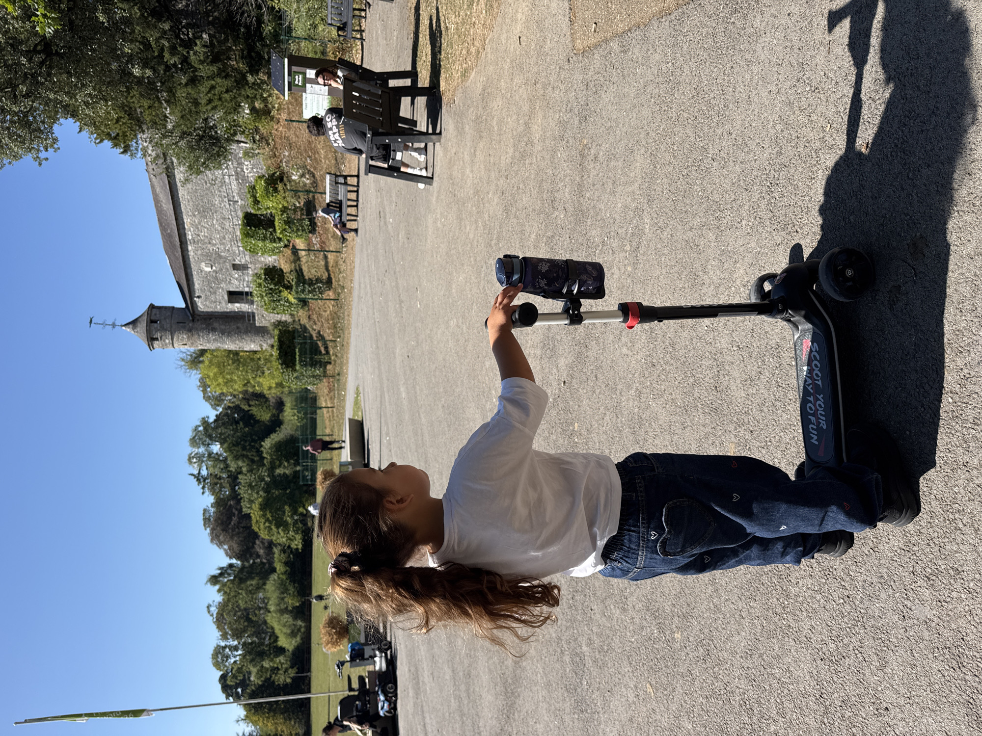 The image depicts a young girl with long, brown hair in a high ponytail, wearing a white t-shirt and dark blue jeans adorned with small white and red hearts. She is standing on a paved pathway, facing away from the camera and to her right, holding onto the handlebar of a black scooter. The scooter has a water bottle attached to its handlebar.
In the background, a stone building with a tower is visible, situated on a hill. This building is Cahir Castle, located in Cahir, Ireland. Beyond the castle, there are trees and a clear blue sky. Several other people are present in the scene, seated on benches in the middle ground, and some walking in the distance. The environment appears to be a park or recreational area, with a flag pole to the left. The sun is shining, casting shadows on the ground.