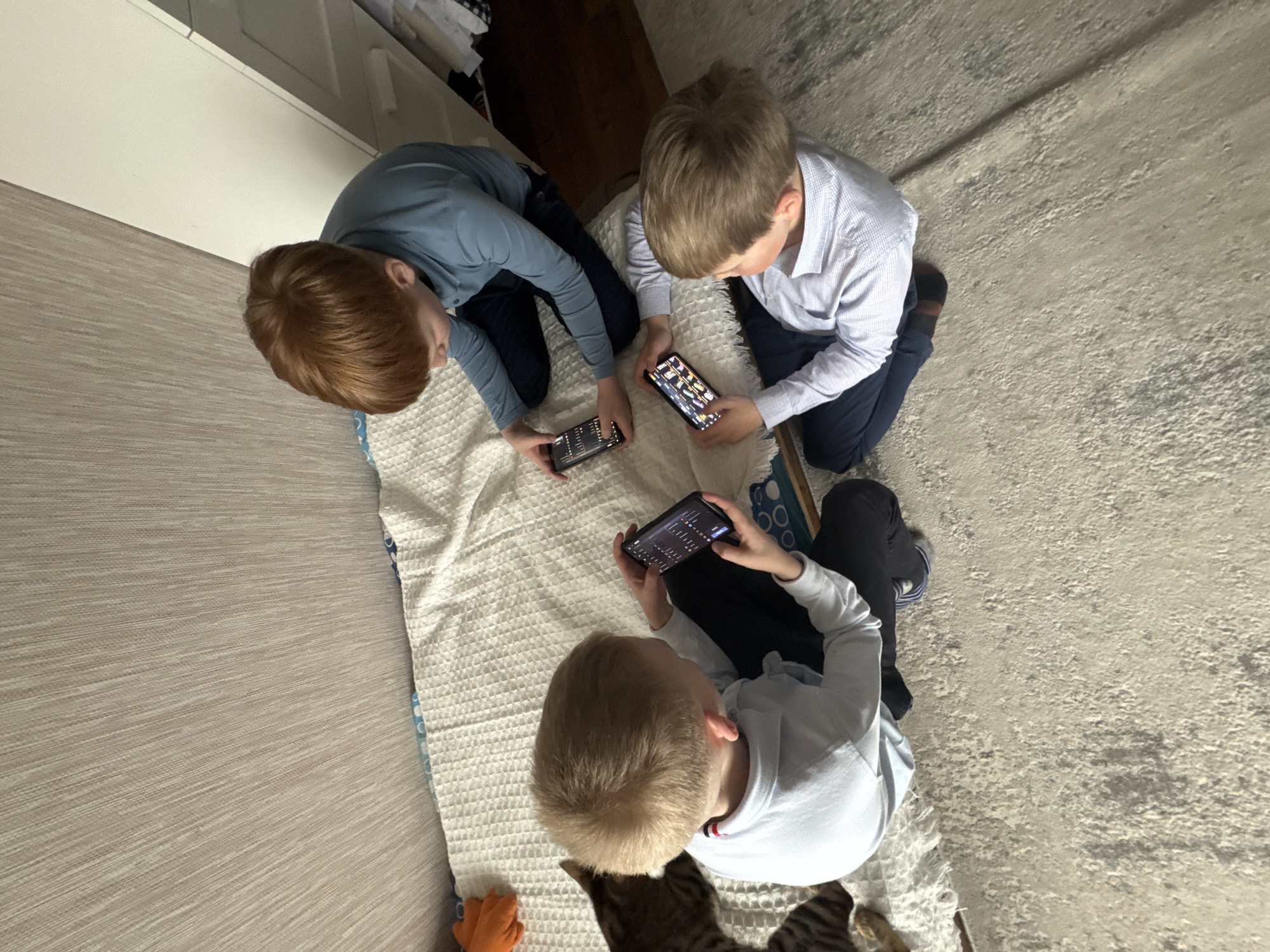Three young boys are kneeling on a blanket placed on the floor. Each boy is holding and looking at a mobile phone. The room is indoors and appears to be a domestic setting. The floor is carpeted with a neutral color. A white cabinet is visible along the wall. The image was not taken in Votkinsk, Russia.