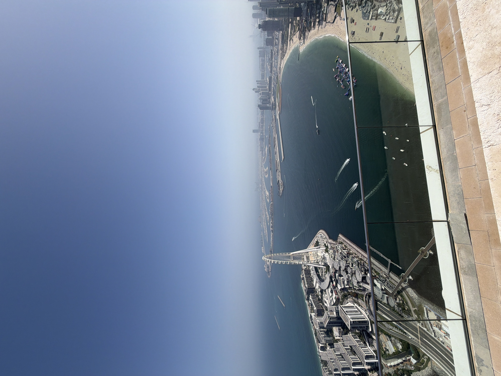 The image shows an aerial view of a portion of Dubai, United Arab Emirates, taken from a high vantage point, likely a skyscraper. To the right, there is a partial view of a window or a glass barrier, suggesting the photo was taken from inside a building. A large body of water, presumably the Arabian Gulf, dominates the central part of the scene. The Palm Jumeirah is visible, a man-made archipelago shaped like a palm tree. Various boats are scattered across the water, indicating maritime activity. The Dubai Marina skyline can be seen in the distance, characterized by numerous high-rise buildings. The landscape transitions from the urban environment to the clear, blue sky.
