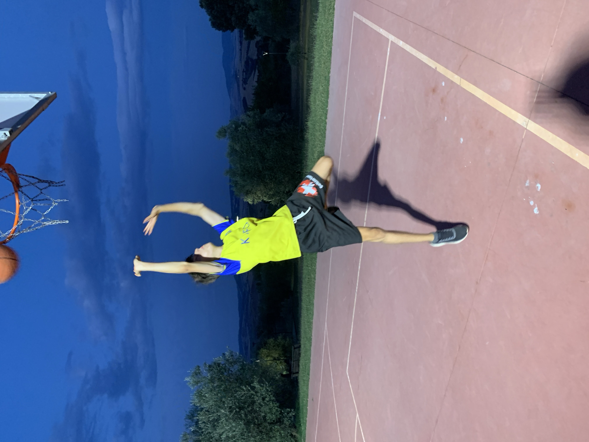 A young person is captured mid-air, attempting a basketball shot at dusk on an outdoor court. The subject, wearing a bright yellow and blue t-shirt and black shorts, is positioned towards the right of the frame, with their arms extended upwards towards the hoop. A basketball is visible in the upper portion of the image, near the net. The basketball court, with its distinctive red-brown surface and white and yellow markings, occupies the foreground and mid-ground. The background reveals a gently rolling landscape with scattered trees and hills, indicative of a rural setting. The sky above is a deep blue, with visible clouds, suggesting the transition from day to night. The location is identified as an outdoor basketball court in San Quirico d'Orcia, Italy.