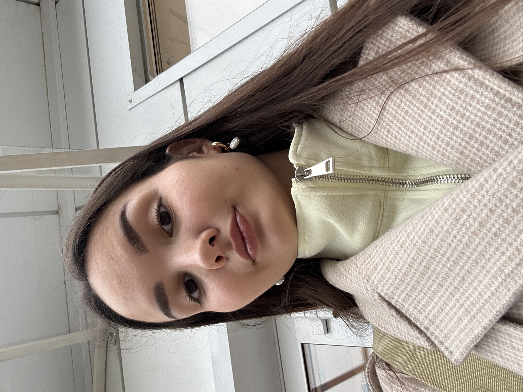 A woman with long, dark hair and light skin is wearing a tan and white houndstooth coat over a light yellow zippered top. She is looking directly at the camera. She is wearing pearl earrings in each ear. The background appears to be an exterior wall of a building with a window and some pipes or ductwork. Location: Chaikovsky, Russia.