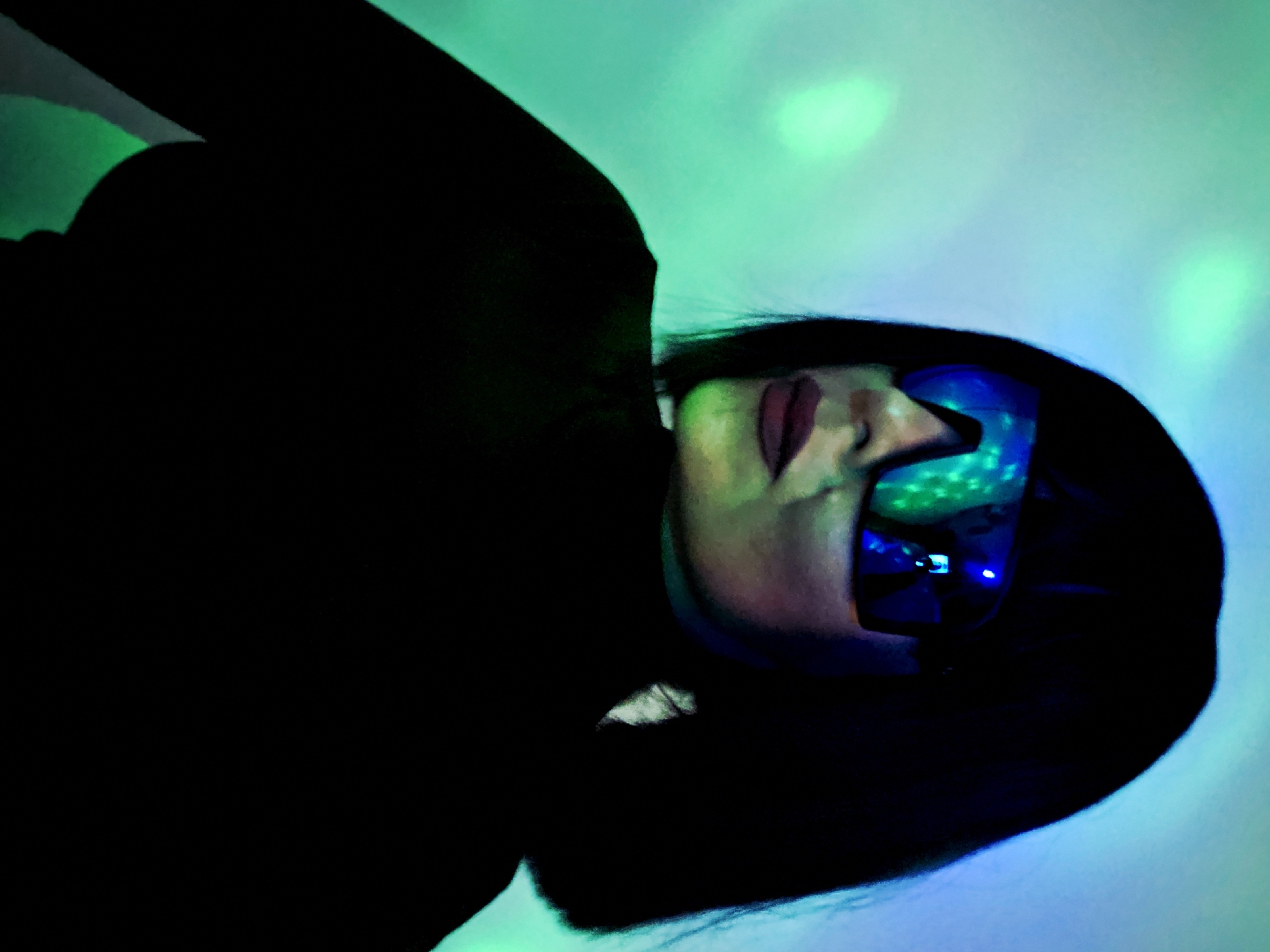 A woman wearing sunglasses is positioned against a green and blue illuminated background. Her dark hair frames her face, and she is wearing dark clothing. The environment is dark.