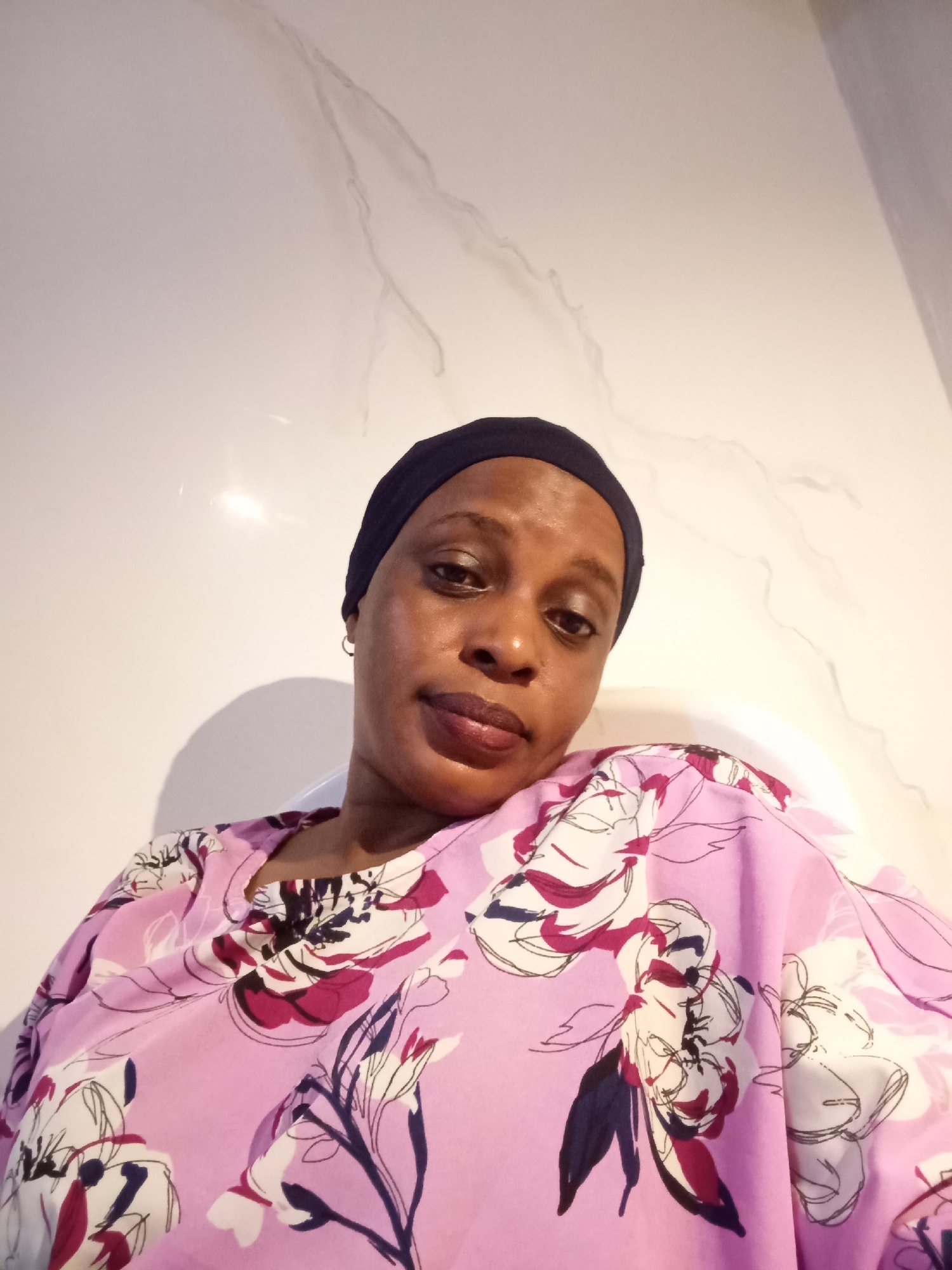 A woman with dark skin and a black head covering is the subject of a selfie. She wears a floral-patterned pink top. The background is a white wall with a faint marble-like design. The location is presumed to be in Unguwar Rimi, Nigeria.