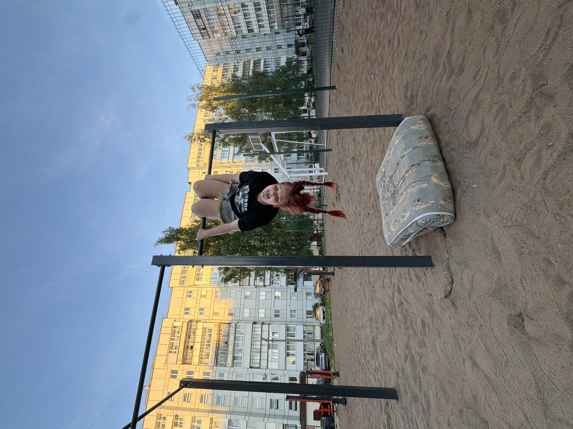 The image depicts an outdoor exercise area, likely in a residential neighborhood in Chaikovsky, Russia, during what appears to be late afternoon or early evening. A young person, with reddish-brown hair styled in two long braids, is the central subject. She is actively engaged in a playful maneuver, hanging upside down from a black metal pull-up bar. She wears a black t-shirt with an unidentifiable graphic design and light-colored shorts, and her expression is jovial, with her tongue sticking out.
Adjacent to the pull-up bar, a worn, light blue mattress with a distinct pattern of small figures or animals is placed on the ground, seemingly as a safety measure. The ground of the exercise area is covered in light brown sand, showing various textures and imprints.
In the background, several large, multi-story residential buildings, characteristic of Soviet or post-Soviet architecture, are visible under a clear blue sky. Some buildings reflect a warm, yellowish light, while others appear cooler in tone. Green foliage from trees is interspersed among the buildings. A metal fence separates the immediate playground area from what appears to be a parking or service road, with a few cars partially visible. Other exercise equipment and a basketball hoop are also present further in the background. No discernible text is clearly visible on any object.