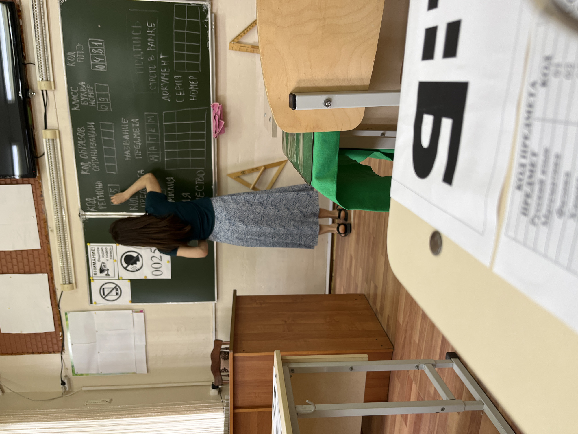 This image captures the interior of a classroom in Kaspiysk, Russia, focused on a female figure writing on a blackboard. The classroom features light-colored walls, a wooden floor, and artificial overhead lighting from two long fixtures above the blackboard. Natural light enters from a window on the left, partially obscured by vertical blinds.
On the left side of the room, a wooden teacher's desk and chair are visible. In the foreground, a light-colored student desk holds several papers. One paper prominently displays a large black Cyrillic letter "Я:" (Ya:). Another paper below it shows a table listing subjects like "Русский язык" (Russian language), "Математика" (Mathematics), "Физика" (Physics), and "Химия" (Chemistry) with corresponding codes "01", "02", "03", and "04". A dark coin rests on this desk.
The primary subject is a female individual, seen from behind, standing in front of a large green blackboard. She wears a dark green top, a long patterned skirt (grey and white tones), and dark sandals. Her long dark hair is down. She is actively writing on the blackboard with chalk in her right hand.
The blackboard is meticulously formatted with chalk-drawn boxes and text. Visible headings include: "КОД РЕГИОНА" (REGION CODE) with "15" entered, "КОД ОБРАЗОВ ОРГАНИЗАЦИИ" (EDUCATIONAL ORGANIZATION CODE), "КЛАСС БУКВА НОМЕР" (CLASS LETTER NUMBER) with "09" entered, and "КОД ППЭ" (P.P.E. Code, likely Exam Conducting Point Code) with "0481" entered. Below these, "НАЗВАНИЕ ПРЕДМЕТА" (SUBJECT NAME) has "МАТЕМ" (likely truncated "Математика" for Mathematics) written in a box. Further right, "ПОДПИСЬ СТРОГО В РАМКЕ" (SIGNATURE STRICTLY WITHIN THE FRAME) and "ДОКУМЕНТ СЕРИЯ НОМЕР" (DOCUMENT SERIES NUMBER) are visible. A small pink cloth hangs on the bottom edge of the blackboard.
Mounted on the wall to the left of the blackboard are three signs: a "ВНИМАНИЕ!" (ATTENTION!) sign stating "Ведётся аудио и видео запись" (Audio and video recording is taking place), a "no phones" icon, and a "no talking" icon, below which are the numbers "0025". A flat-screen television is mounted high above the blackboard, and a long, horizontal heater or radiator is affixed to the wall below the TV. In the midground, various student desks and chairs are arranged, with one desk covered by a green tablecloth. A wooden set square is leaning against the wall on the right.