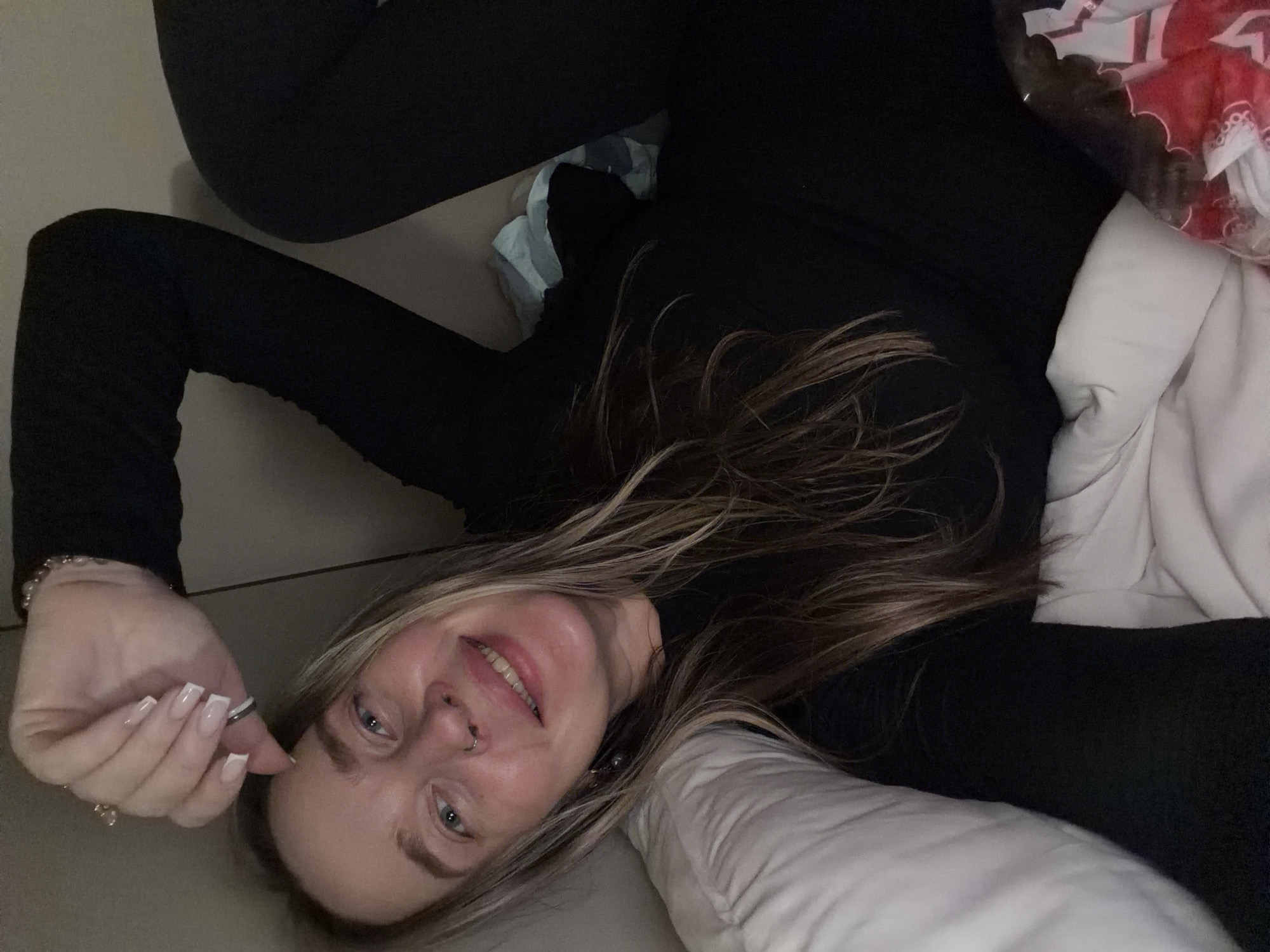 A woman is lying down, seemingly in a domestic setting. She is wearing black leggings and a long-sleeved black top. Her hair is blonde and brown. She has a nose piercing on the left side. Her right hand is visible, with manicured nails. Part of a white pillow is visible near her head and torso, suggesting she might be in bed or on a couch. Red and white patterned fabric is visible in the background. The location is unknown.