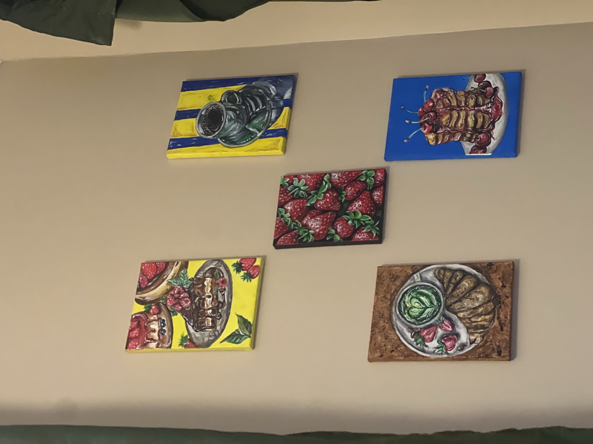 The image displays a collection of five small, colorful paintings of food items hanging on a plain, beige wall. The paintings are arranged in a staggered formation: two paintings are on the upper left and upper right, one painting is in the center, and two paintings are on the lower left and lower right.
The painting on the upper left depicts a slice of chocolate cake topped with strawberries and blueberries on a yellow background. To its right, a painting on the upper right shows a cup of coffee in a dark cup and saucer against a background of blue and yellow stripes. The center painting features a close-up of ripe strawberries on a black background. The painting on the lower left illustrates a croissant with a cup of matcha latte, adorned with a heart shape, and three strawberries on a light-colored plate, set against a textured brown background. Finally, the painting on the lower right presents a stack of pancakes topped with cherries and syrup on a white plate, against a vibrant blue background.
The wall appears to be indoors, with a portion of a dark green curtain visible on the far left and a glimpse of a dark green garment or object on the upper right. The overall scene is domestic, possibly a kitchen or dining area. There are no people visible in the image. The lighting is soft and even, suggesting a well-lit interior space.
The location in Varna, Bulgaria, is not ascertainable from the provided image.
