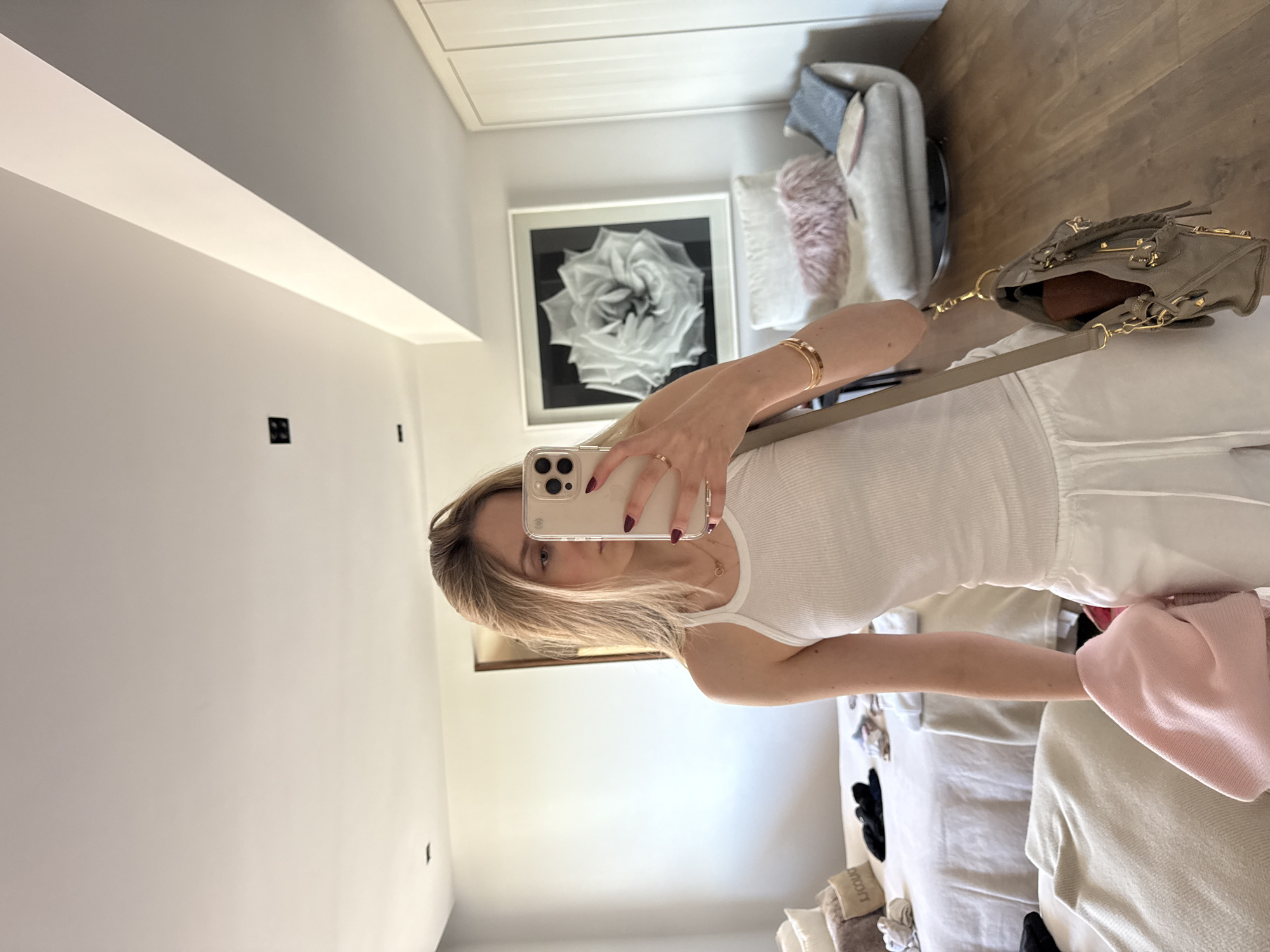 A young adult female, with long blonde hair partially covering her face, is depicted in a full-length mirror selfie inside a brightly lit room. She is wearing a white ribbed tank top, white drawstring pants, two delicate gold necklaces, and two gold bangles on her left wrist. Her fingernails are painted dark red. She holds a silver smartphone with a clear case in her right hand, angled towards the mirror to capture the image. Her left hand holds a light pink knitted garment. A grey shoulder bag with prominent gold hardware hangs from her left shoulder, revealing a brown object within its open top.
The room features light wooden floorboards and white walls. To the left, a bed with white bedding is visible, holding multiple pillows, including one with "JACQUEMUS" partially legible, and various clothing items like black sandals. To the right, a white swivel armchair with a pink faux fur pillow and a blue throw pillow sits beneath a large framed black and white photograph of a rose. A tall, white two-door wardrobe is located further right. The white ceiling has a visible beam structure and a single black square recessed light fixture. There are no identifiable elements within the image to confirm the location as Mexico City, Mexico.
