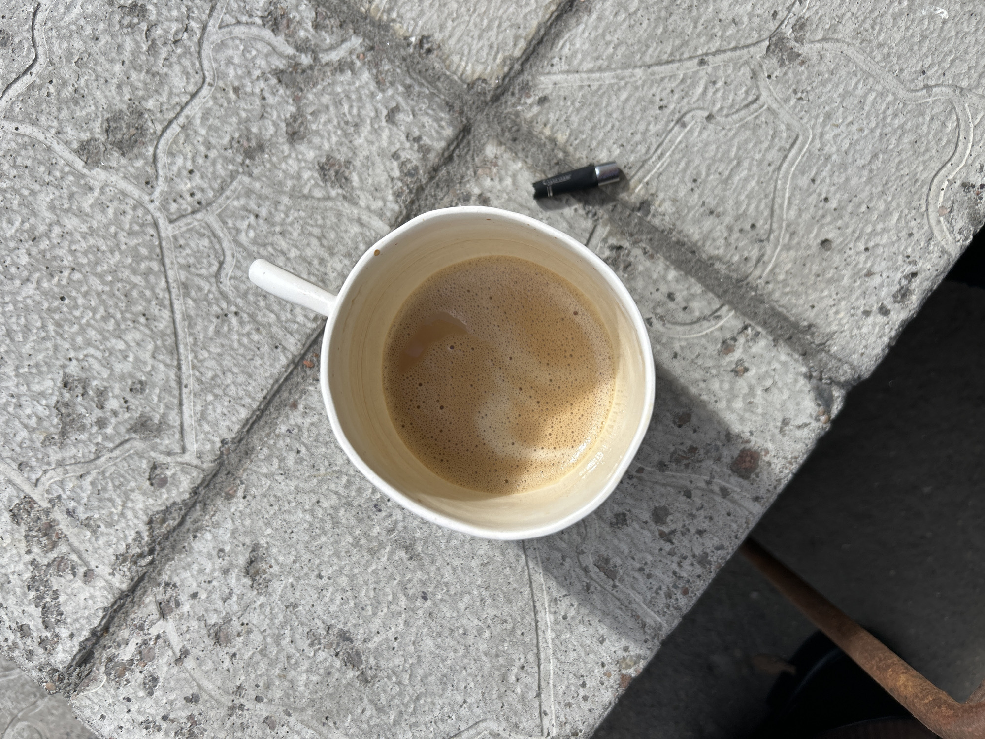 An overhead shot presents a white ceramic mug, filled with a light brown, foamy beverage, likely coffee, resting on a textured, light gray concrete surface. Bright sunlight illuminates the scene, creating sharp, distinct shadows, particularly a large one that covers the lower right corner and reveals a dark, possibly metallic, bar.
The concrete surface is rough and mottled with small dark speckles. It features intricate, shallowly carved or etched patterns resembling swirling lines and branching structures, which are lighter in color. Visible seams and cracks run through the concrete.
Near the top right edge of the mug, a small black cylindrical object with a silver end is present. The text "Clipper" is clearly visible on this object, identifying it as a lighter. The presence of the mug suggests a moment of rest or a break, possibly outdoors on a patio or ledge, under a sunny sky. No people or active events are depicted.
