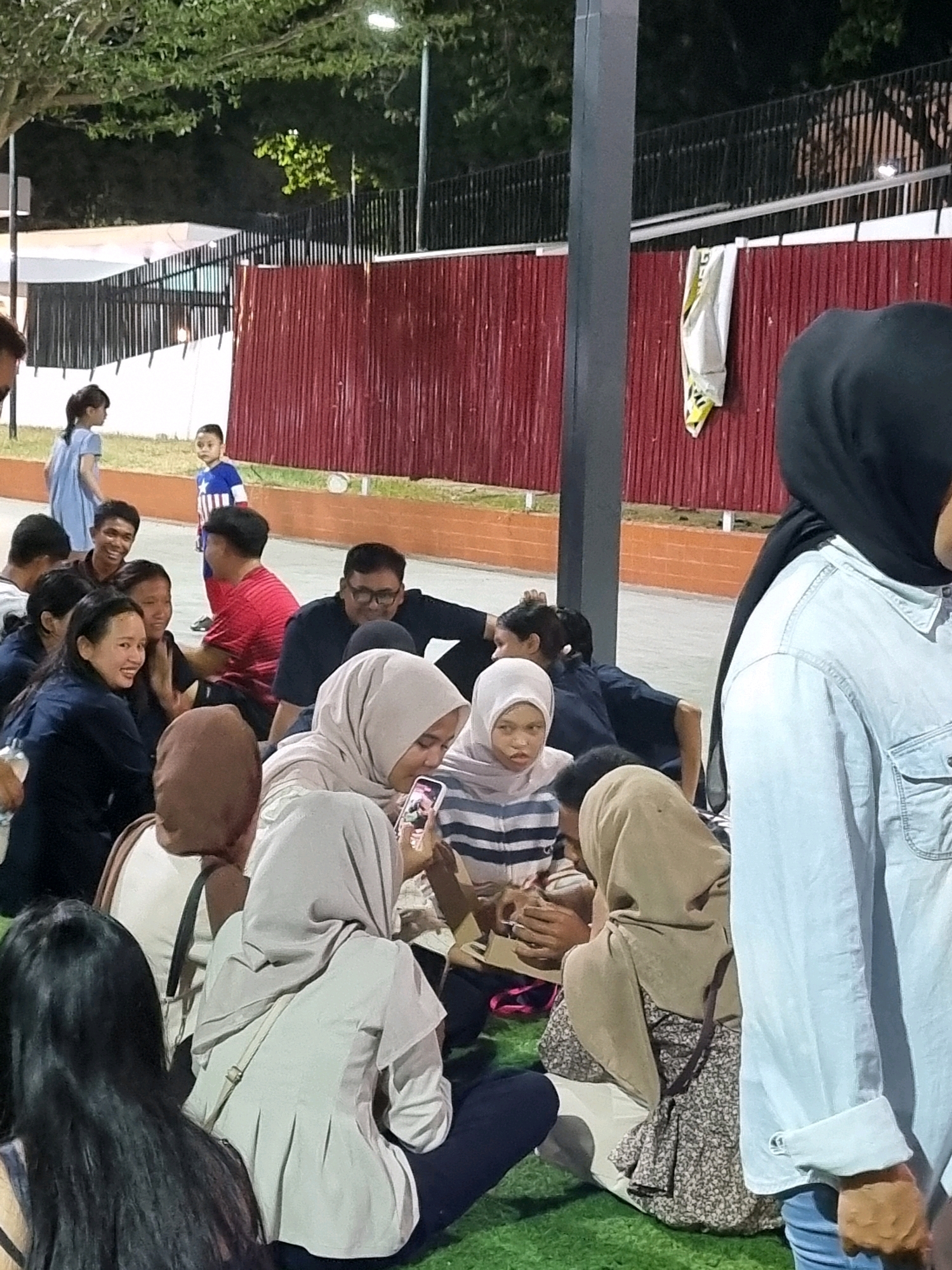 In Tanamodindi, Indonesia, at dusk, a group of young people are gathered on a grassy area in front of a dark red corrugated metal fence. Some individuals are sitting on the ground, while others appear to be standing. A tall, dark grey pole stands prominently in the foreground, casting a shadow. In the background, trees with green foliage are visible, and streetlights illuminate the scene. Several individuals are interacting with each other, some looking at phones. Two children are also present in the background, one standing and looking towards the group, and another in a blue shirt with a star pattern. The atmosphere appears casual and social.