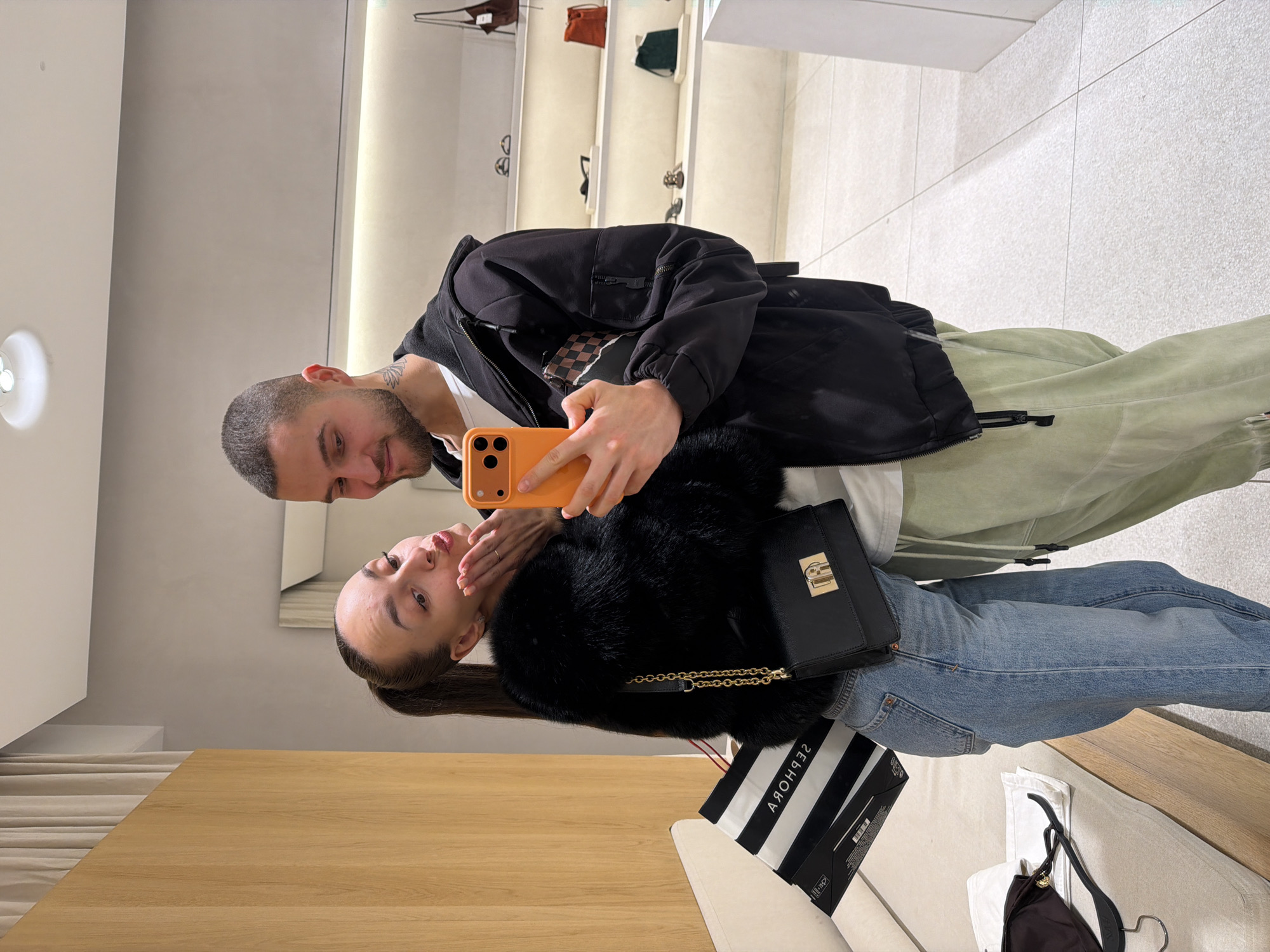 A male and a female are depicted in a mirror selfie taken within a modern retail environment, likely a shopping mall in Sofia, Bulgaria. The male, with short dark hair, a beard, and a neck tattoo, holds an orange smartphone to capture the image. He wears a black hooded jacket over a white t-shirt and light green pants, and is smiling subtly. The female, positioned to his left, has dark hair in a ponytail and is wearing a black faux fur jacket, blue jeans, and a small black shoulder bag with gold hardware. She poses with puckered lips and cupped hands directed towards the phone. A reflected "Sephora" shopping bag rests on a light-colored bench or seating area to her left. The background features minimalist white shelves displaying various handbags and shoes, illuminated by recessed lighting. The walls are a combination of light grey paint and light wood paneling, complementing the light-colored tiled floor.