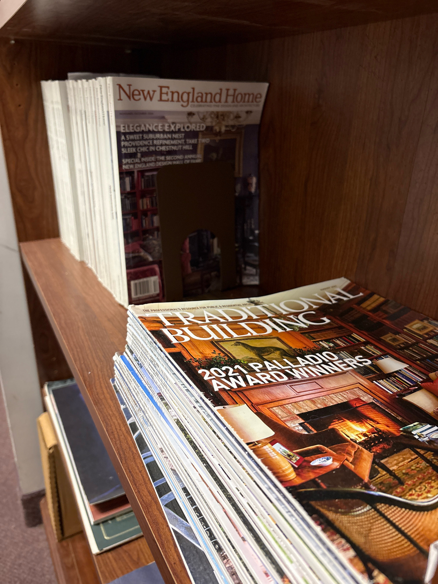 The image displays a bookshelf in what appears to be a residential or small commercial setting in Windsor, United States of America. The bookshelf is made of dark wood and contains several magazines. On the top shelf, a stack of "New England Home" magazines is visible. The covers show text such as "Elegance Explored," "A Sweet Suburban Nest," and "Providence Refinement." Below this, on the middle shelf, a stack of "Traditional Building" magazines is prominently displayed, with the headline "2021 Palladio Award Winners" clearly visible on the top magazine. The interior of the room is not fully visible, but the lighting suggests it is indoors.