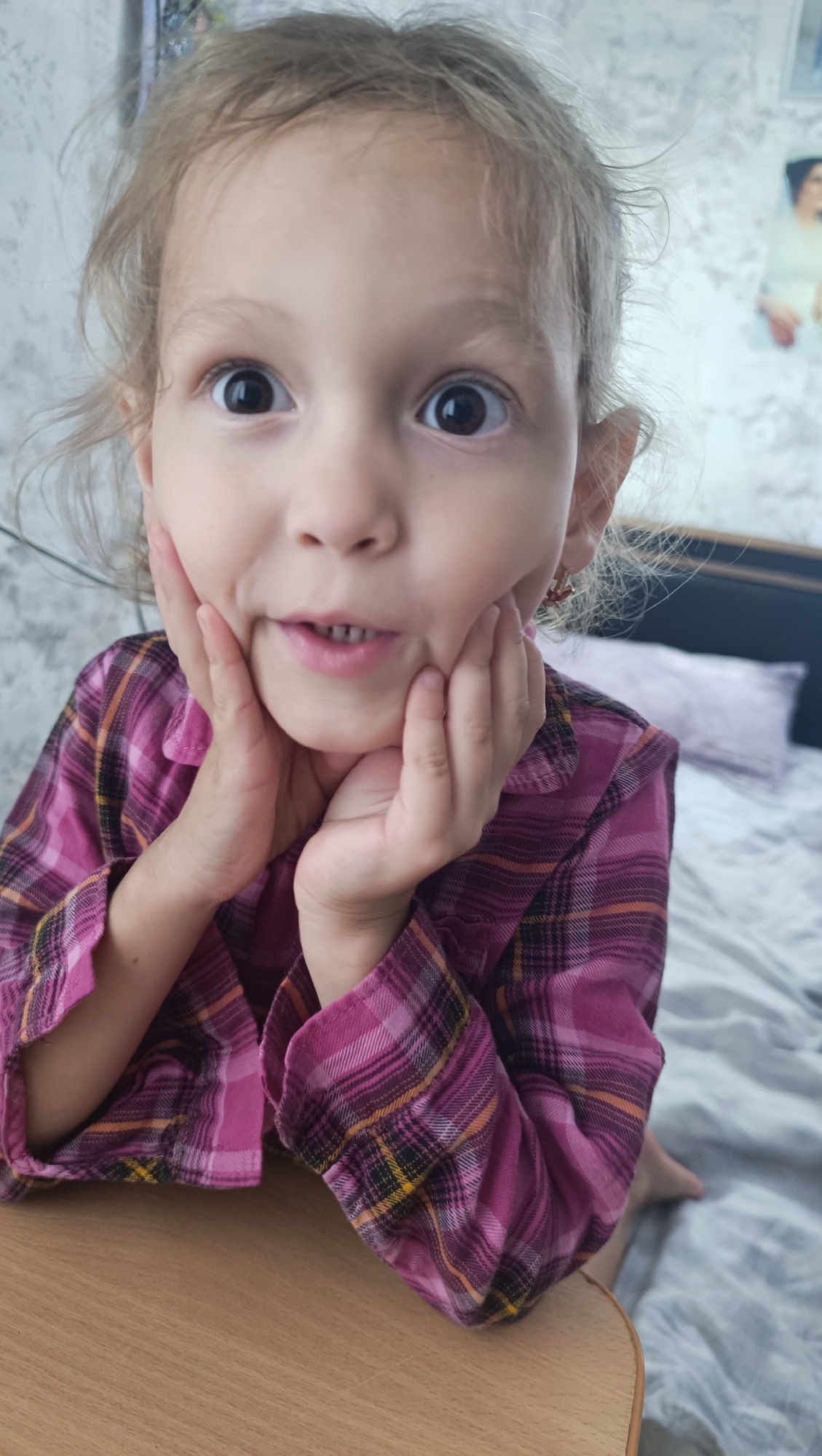 A young girl with light brown hair is leaning forward, resting her chin on her hands. Her eyes are wide and dark, and her mouth is slightly open, revealing her teeth. She is wearing a pink and purple plaid shirt. She is positioned in front of a wooden surface, likely a table. The background appears to be a bedroom with a bed and a wall with a framed picture. The scene is well-lit. There is no indication of the city of Karsha, Russia in the image.