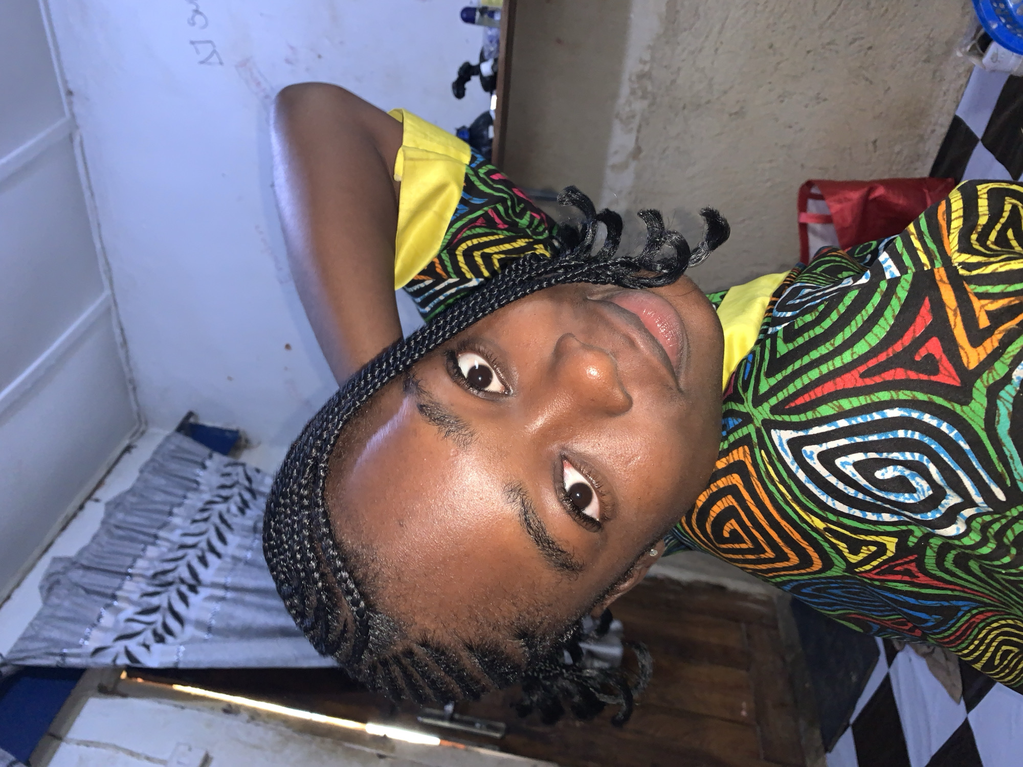 The image shows a close-up of a young Black female with braided hair, wearing a brightly colored, patterned shirt with a yellow collar. She is looking at the camera with her head tilted and her right arm behind her head. The background is a room with a light blue wall, a shelf with items on it, and a checkered floor. There is also a red bag visible in the background. The overall setting appears to be indoors, possibly a residential space. The image does not provide any specific visual cues to determine the exact location within Rafin Par, Nigeria.