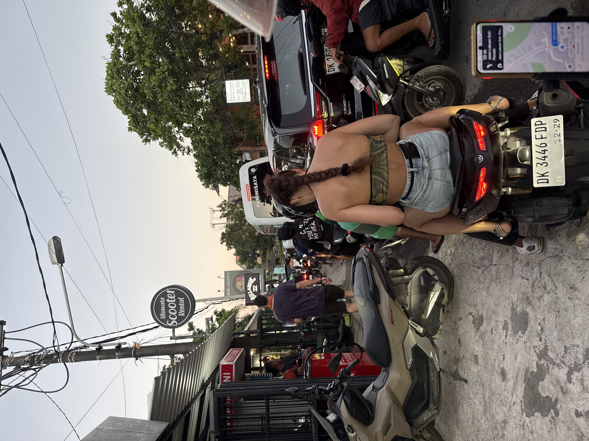 A congested street scene in Kuta Selatan, Indonesia, depicts a woman with a long braid riding pillion on a motor scooter, revealing her bare back. Adjacent to her, another scooter is positioned, its rider navigating with a smartphone showing a map app. The immediate area teems with parked and operating scooters, creating a dense traffic situation. Signage for 'Uluwatu Scooter Rental' and 'Kitchen' is visible. The street also features shops, pedestrians, utility poles laden with wires, and various motor vehicles, all under a hazy sky.