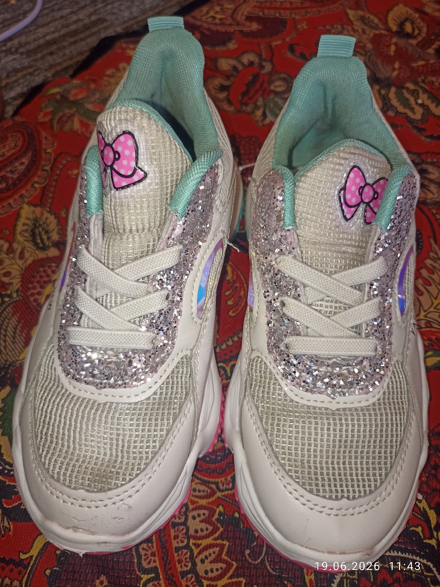 A pair of white and pink children's sneakers with teal accents and glitter embellishments are displayed on a red and gold patterned fabric. Each shoe features a pink bow graphic with white polka dots on the heel. The sneakers have white laces and mesh detailing on the front. The background fabric has a paisley-like pattern. The lighting suggests an indoor setting, and a timestamp "19.06.2026 11:43" is overlaid on the bottom right of the image.