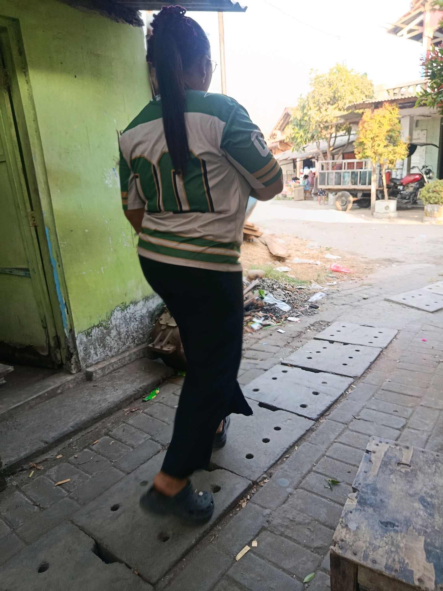 A person walks past a light green building on a street in Jobokuto, Indonesia. The person is wearing a green and white striped shirt and black pants, with dark shoes. They are facing away from the camera, walking on a sidewalk composed of concrete slabs and brick pavers. In the background, there are buildings, trees, a truck, and a red motorcycle. The scene is illuminated by daylight.