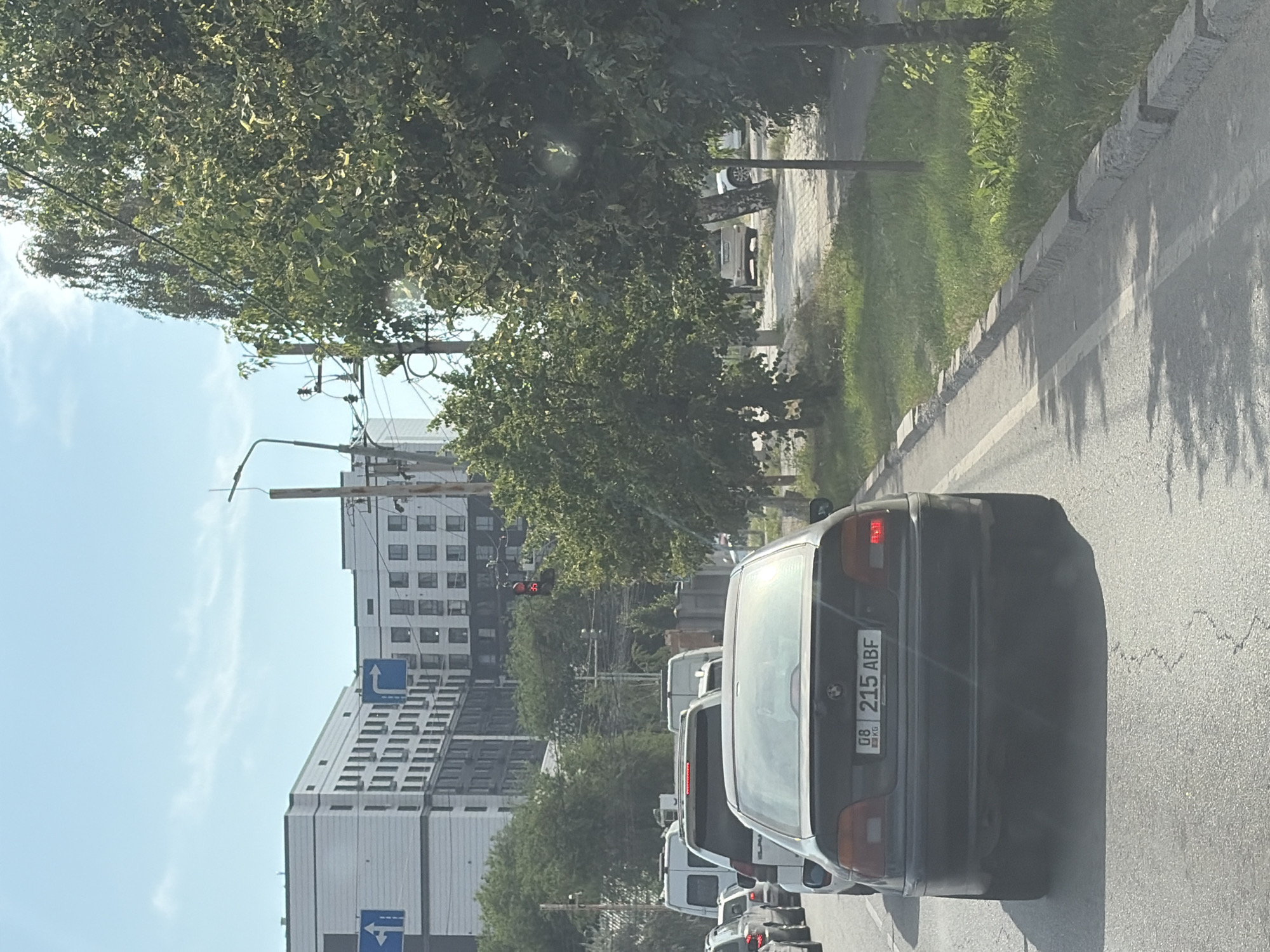 An over-the-shoulder rear view of a black BMW sedan is captured as it travels down a paved road. The car's license plate is visible, reading "08 KG 215 ABF". Other vehicles, including vans and cars, are seen ahead in traffic. The road is flanked by a sidewalk and a grassy area with trees. In the background, modern multi-story buildings and a traffic light are visible. The sky is blue with scattered clouds.