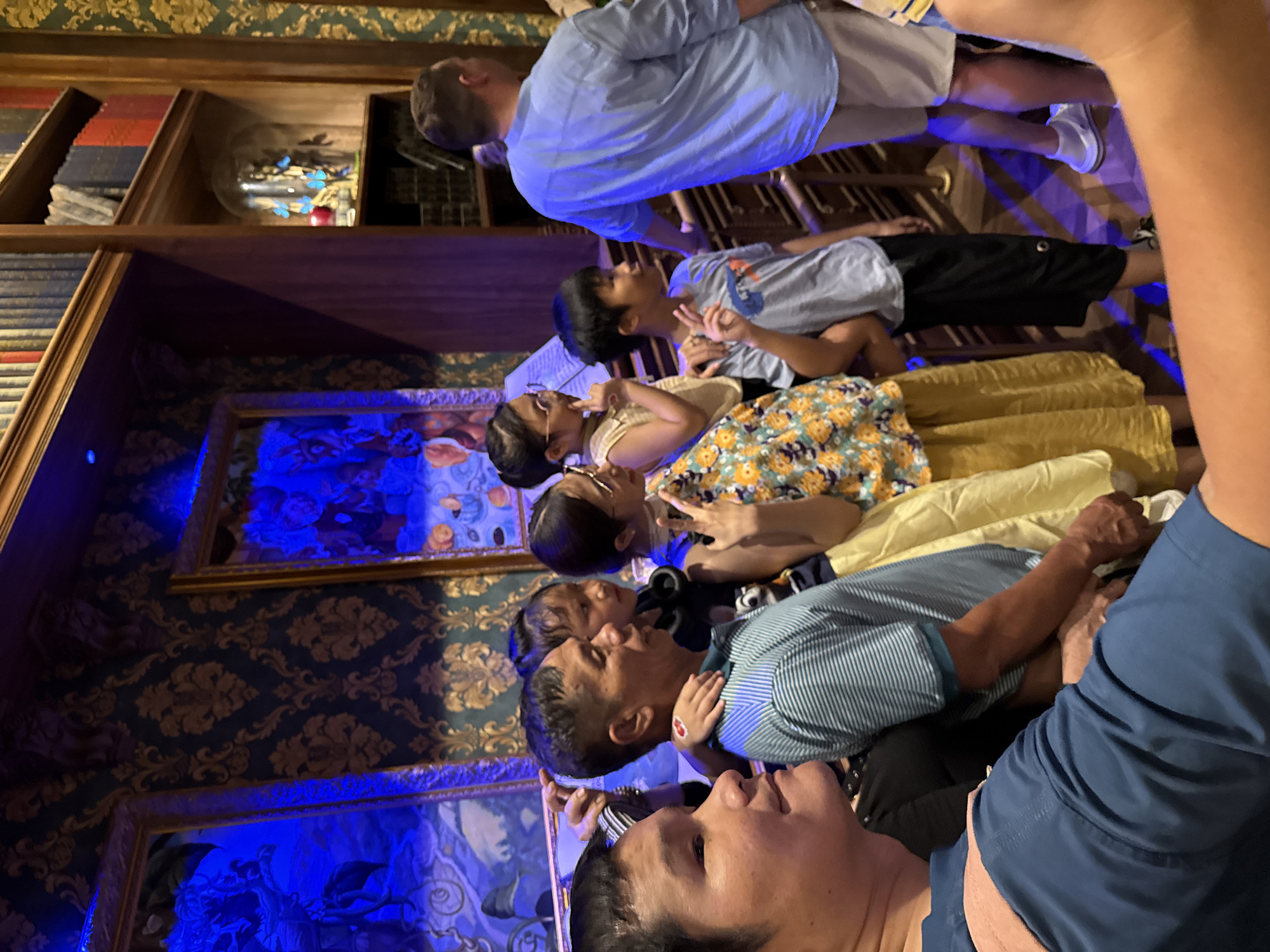This indoor photograph captures a diverse group of people, including adults and children, gathered in a dimly lit, ornately decorated room. All individuals are looking upwards, suggesting they are observing a specific display or event above them.
The setting features elaborate dark wallpaper with a gold and blue damask pattern. To the left, dark wooden bookshelves are visible, filled with numerous books. Along the walls, several framed artworks or displays are prominently illuminated by strong blue light, creating a dramatic and ethereal effect. One particular shelf showcases a glass cloche containing decorative items, possibly butterflies, also glowing with the same blue light.
The subjects are dressed casually. In the foreground, an adult male with short dark hair, wearing a light blue striped polo shirt, looks up. Another adult male in a light blue t-shirt and shorts stands further back, similarly focused upwards. Among the children, a boy in a light blue t-shirt is seen holding up a peace sign, gazing intently. A girl with glasses and dark hair appears thoughtful, with a finger near her mouth, also looking up at the illuminated displays. The collective posture and gaze indicate engagement with whatever is positioned above them.
The blue lighting on the artworks and objects creates a captivating, almost magical atmosphere. The scene evokes a sense of wonder and curiosity among the observers. Given the context of Nha Trang Ward, Vietnam, this location could be a themed attraction, a museum, or an exhibition space designed for immersive experiences. There is no visible legible text in the image.