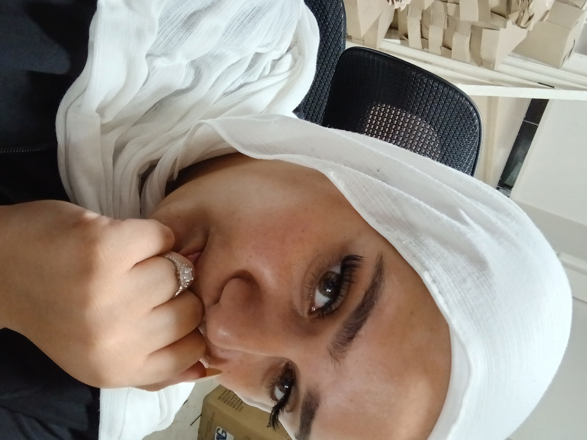 A close-up, eye-level shot focuses on the face of a young woman wearing a white hijab. Her right hand is raised to her mouth, with her fingers gently touching her lips, and she is wearing a ring on her left index finger. The background is blurred, but appears to be an indoor setting with stacks of cardboard boxes and a portion of a black office chair. The woman's eyes are brown, and she has long, dark eyelashes. The lighting is soft, with no harsh shadows. The image is a selfie, taken from the woman's perspective. The location is identified as Rukn ad-Din Municipality, Syria.