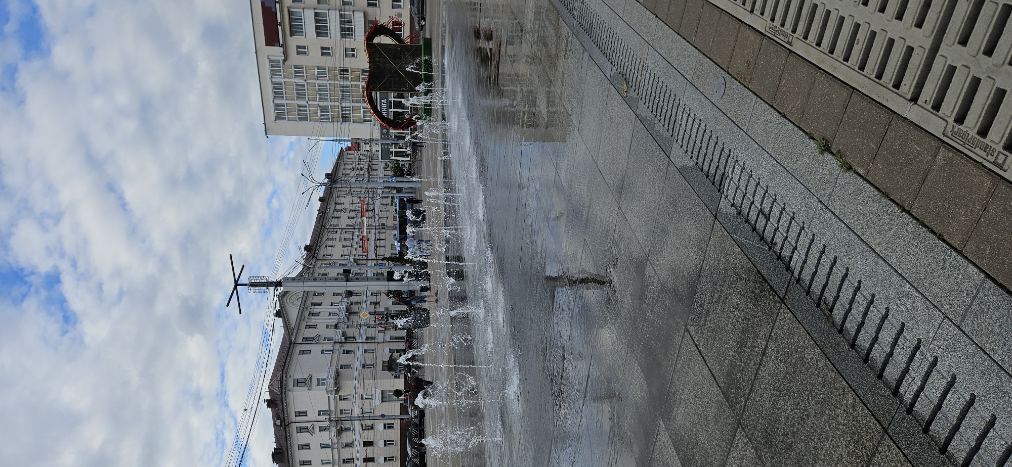 The image depicts a public square in Vitsebsk, Belarus, with an active water fountain in the foreground. The fountain consists of multiple jets of water spraying from the ground, creating a shimmering effect on the wet pavement. The square is paved with large, grey granite tiles. In the background, a line of multi-story buildings with light-colored facades and numerous windows runs along a street. One of the buildings has a prominent sign with Cyrillic lettering, likely indicating a shop or establishment. To the right, a large, stylized, brown sculpture, resembling a cup or bowl with red accents, is visible. People are scattered throughout the square, some walking, others standing near the fountain. The sky is cloudy with patches of blue. The overall scene suggests a lively urban environment on a partly cloudy day.