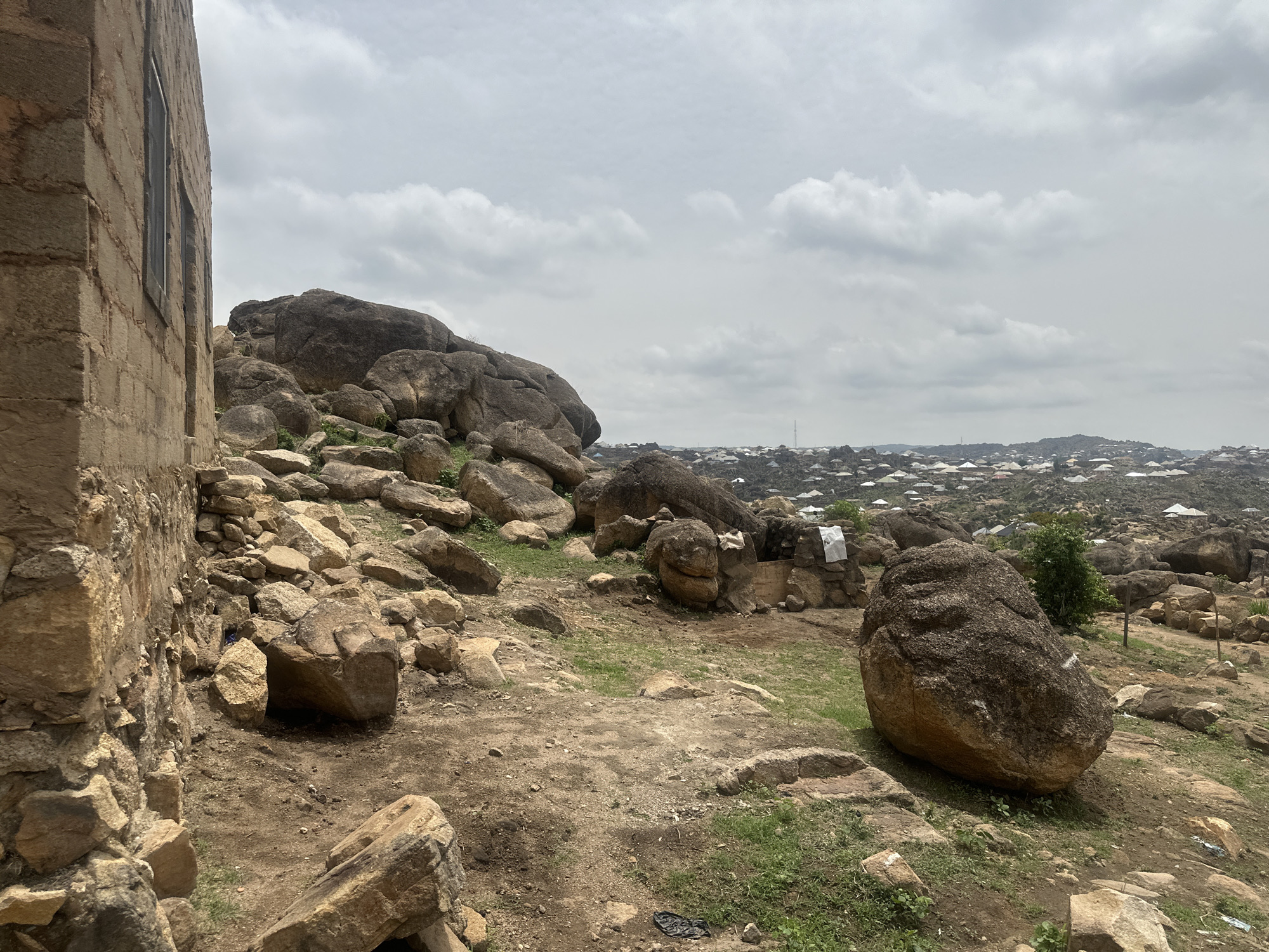 The image depicts a rocky landscape adjacent to a building with stone block walls in the city of Jos, Nigeria. Numerous large, rounded boulders are scattered across the terrain, with sparse patches of vegetation. In the background, a dense urban area with light-colored rooftops can be seen. The sky is overcast with a layer of clouds. The overall setting appears to be on the outskirts of the city, characterized by rocky outcrops and sparse vegetation transitioning into a more densely populated area. The presence of a building suggests a semi-developed area with proximity to both natural rock formations and residential zones.
