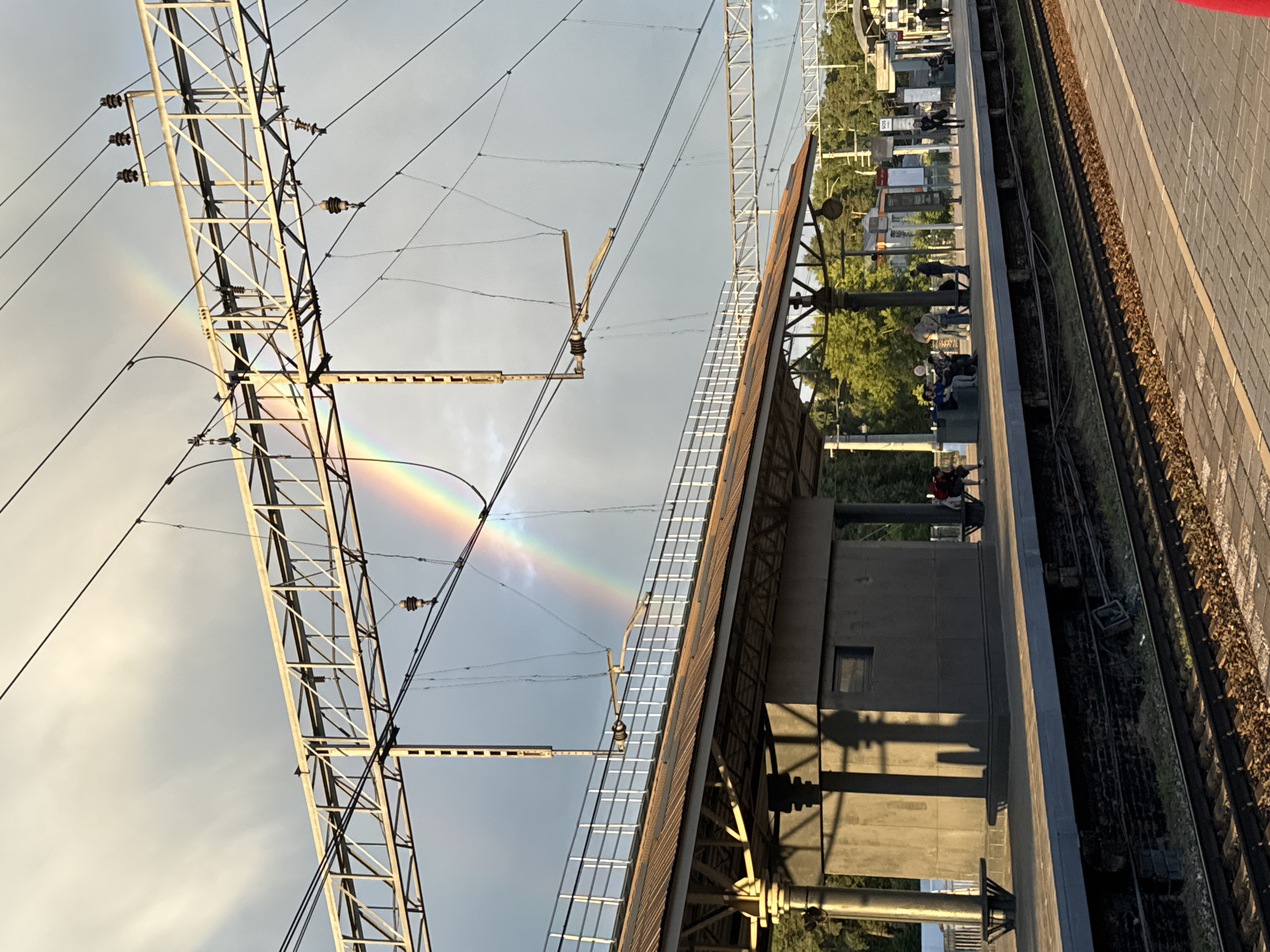 The image depicts a railway platform in Bykovo, Russia, under a cloudy sky where a rainbow is visible. The platform is equipped with overhead electrical wires and support structures for the railway. A covered waiting area with benches and a small building is present on the platform, where several people are seated and standing. Trees and other structures are visible in the background, suggesting an urban or suburban setting. The scene is illuminated by sunlight, casting shadows on the platform and the waiting area. The railway tracks are in the foreground.