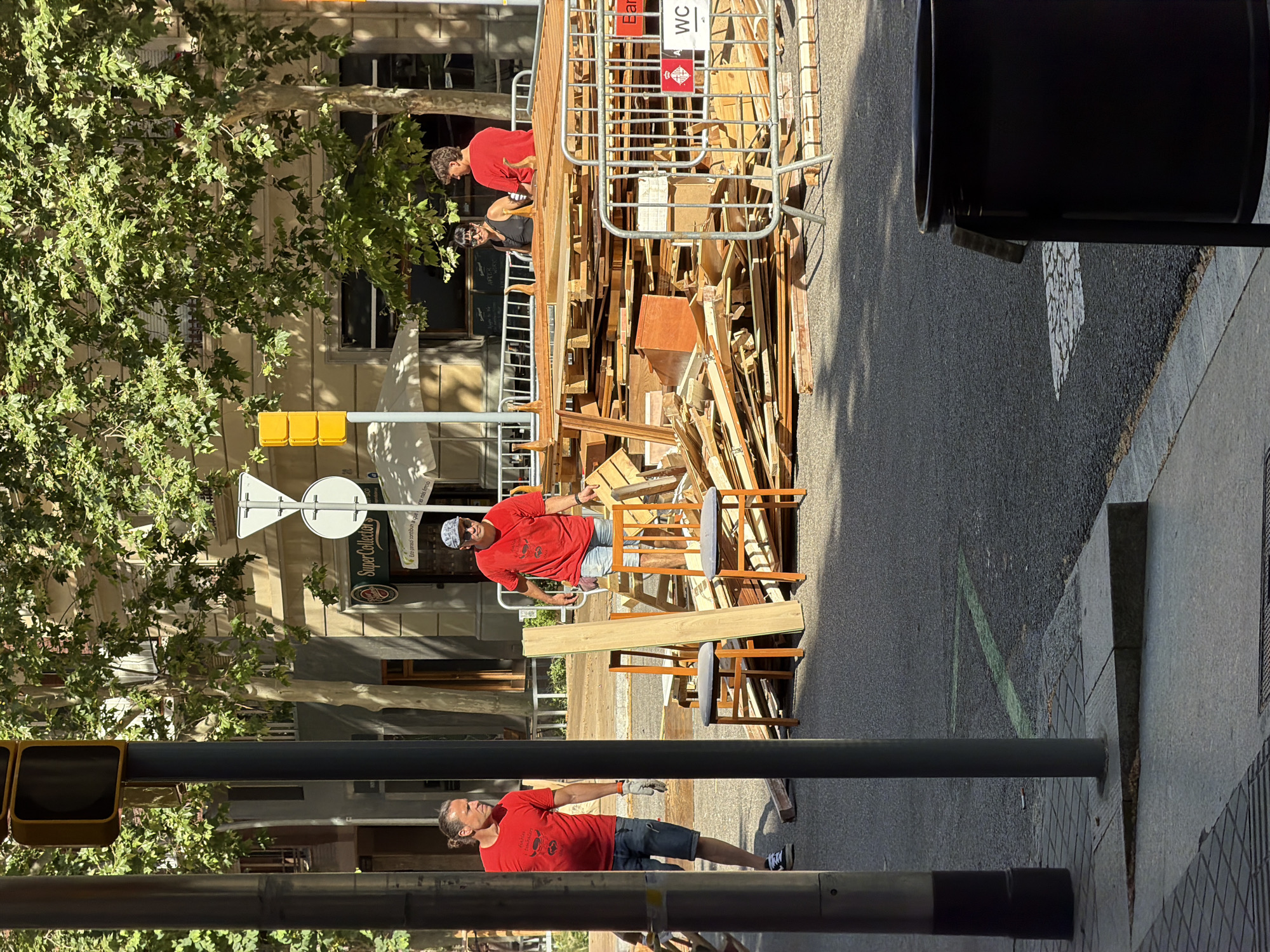 Two men are working on a construction site or debris removal area on a sunny day in Barcelona, Spain. One man, wearing a red t-shirt and denim shorts, is standing and holding a piece of wood. He has long, dark hair and is wearing work gloves. The other man, also in a red t-shirt and denim shorts, is sitting on a chair amongst a pile of wood and construction materials. He is wearing a cap and sunglasses.
The scene appears to be an outdoor urban environment, likely a street or sidewalk, with trees and buildings in the background. A portable toilet with "WC" signage is visible within the construction area. A traffic light and street signs are also present. The overall mood seems to be one of work and manual labor under pleasant weather conditions.