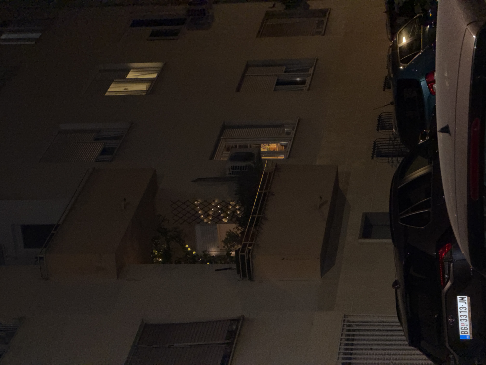 The image shows a nighttime urban scene, likely in the Vracar Urban Municipality, Serbia. The primary subject is the side of a building, showcasing multiple windows with varying degrees of light emanating from within. Some windows are covered with blinds. A balcony decorated with lights and greenery is visible. A parked vehicle, or vehicles, are positioned on the right side of the image, with a license plate clearly showing "BG 3313-JM". The lighting suggests it is either very late in the evening or before dawn. The overall atmosphere is dark.