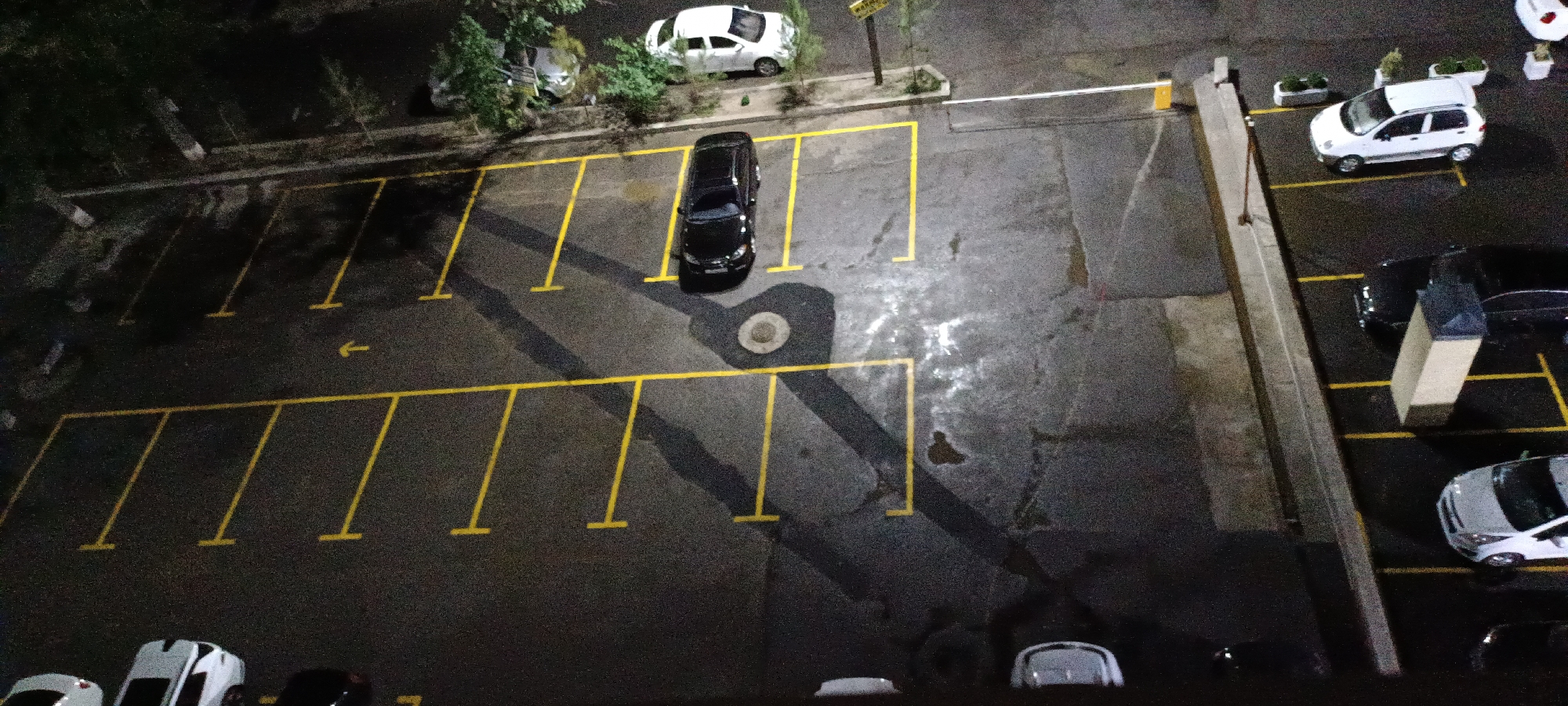 The image shows an aerial view of a parking lot at night, likely in Fergana City, Uzbekistan, given the context. The parking lot is marked with yellow lines indicating parking spaces. Several vehicles are parked in the lot, including a black car in the foreground, a white car on the right, and another white car further up. Trees are visible on the left side of the image, casting shadows on the ground. The lighting suggests artificial illumination from streetlights or building lights. The parking lot appears to be paved with asphalt and has some wet patches. A white barrier arm is visible at the entrance of the parking lot on the right side.