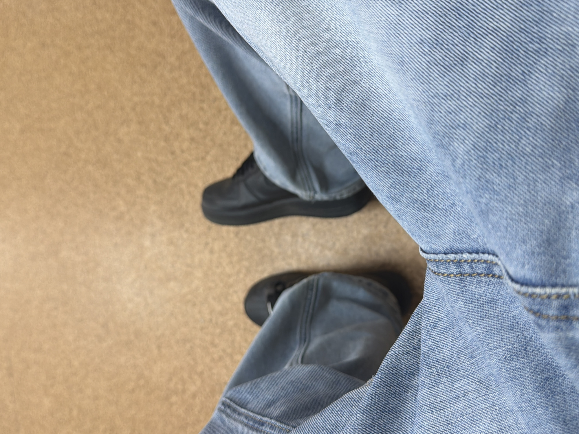 The image shows a close-up view of a person's legs, clad in light blue denim jeans and black shoes, seated on a light brown, textured surface. The perspective is from above, looking down at the legs. One leg is bent at the knee, with the foot flat on the ground. The other leg is slightly extended. The jeans are the primary focus, with visible stitching and the texture of the denim. The shoes appear to be low-top sneakers. The background is a plain, uniform color and texture, suggesting an indoor setting like an office floor or a waiting area. There is no indication of Yekaterinburg, Russia, or any other specific city in the image.