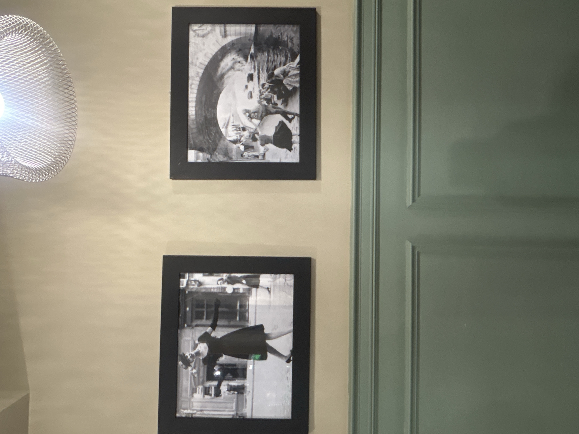 The image shows two framed black and white photographs displayed on a textured cream-colored wall. The wall features a horizontal green wainscoting with decorative molding. Above the wainscoting, the wall has a slight sheen, reflecting some light.
The photograph on the left depicts a woman in a dark, short-sleeved dress and a hat, with elbow-length gloves. She is in motion, with her left leg extended and her right arm outstretched. A small green bag is visible at her hip. In the background, a building with windows and doors is visible, and another person in a dark dress stands to the right.
The photograph on the right shows a scene under a stone archway, likely a bridge. Several people are gathered near a body of water, some sitting on the bank and others in a boat. A woman in a dark, full skirt is seen in the foreground, facing away from the viewer, with her arms raised as if dancing or gesturing.
A metallic, mesh light fixture is visible at the top of the frame, casting a bright light. The overall scene appears to be indoors, possibly in a restaurant, cafe, or gallery. There is no visual information within the image that would allow for the identification of the city of Almaty, Kazakhstan.