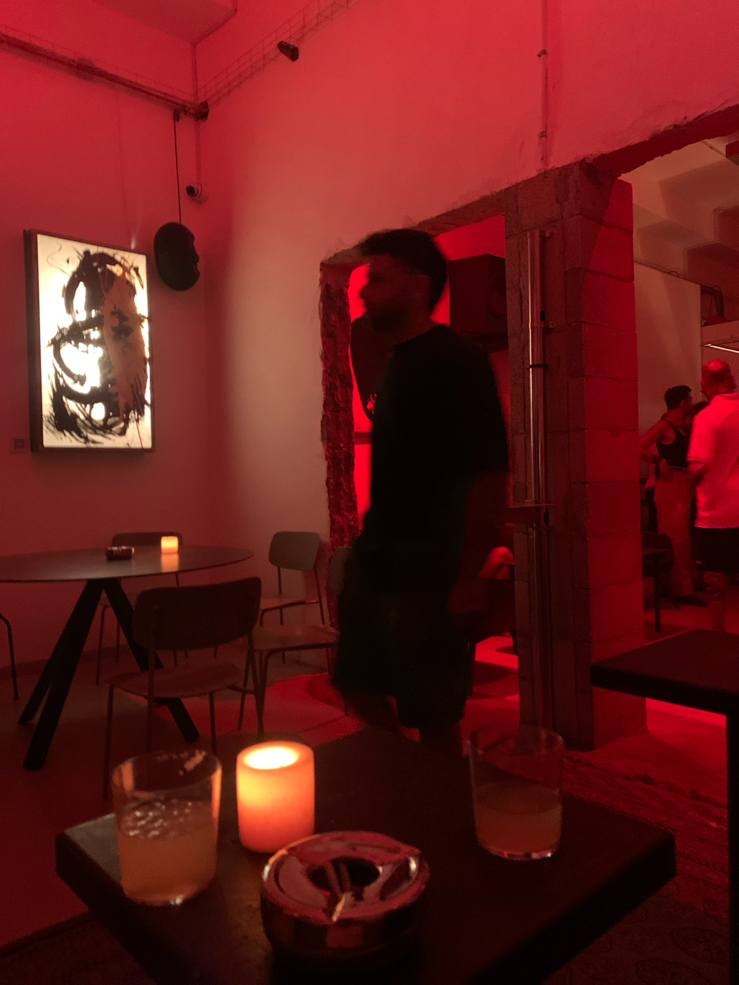 The image depicts an indoor scene illuminated by red lighting, likely a bar or lounge. In the foreground, on a dark table, are two glasses containing a yellow beverage, an ashtray, and a lit candle. In the midground, a man in a dark shirt is visible, walking away from the camera. Further back, several other individuals are present, some interacting with each other. A large piece of abstract art hangs on the wall to the left. The environment suggests a social gathering or nightlife setting. The presence of specific architectural elements and the overall ambiance may indicate a location in Tel Aviv, Israel, known for its vibrant nightlife and arts scene.