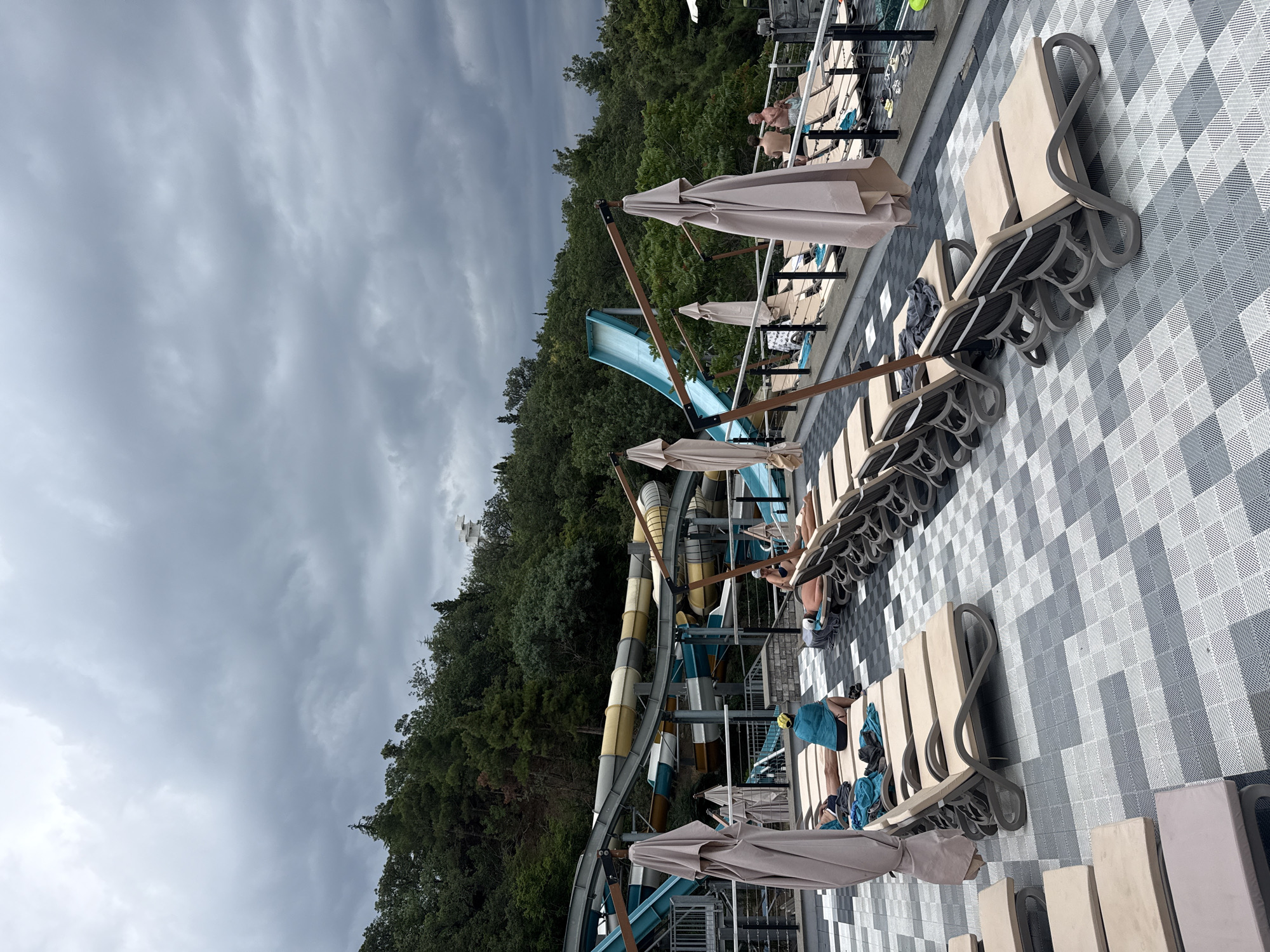 This image depicts an outdoor area, likely at a water park, with numerous lounge chairs and umbrellas arranged on a checkered grey and white tiled surface. In the background, a large water slide structure with yellow, white, and blue sections is visible amidst a dense green forest. The sky is overcast with dark, heavy clouds. Several people are seen relaxing on the lounge chairs. One person, wearing a yellow head covering, is seated on a chair with a blue towel. Another person is lying on a lounge chair, facing away from the camera, with their back exposed. Further in the background, a couple is conversing while seated on lounge chairs. The overall scene suggests a leisure environment, potentially in the city of Yalta, Ukraine, though this cannot be definitively confirmed from the image alone.