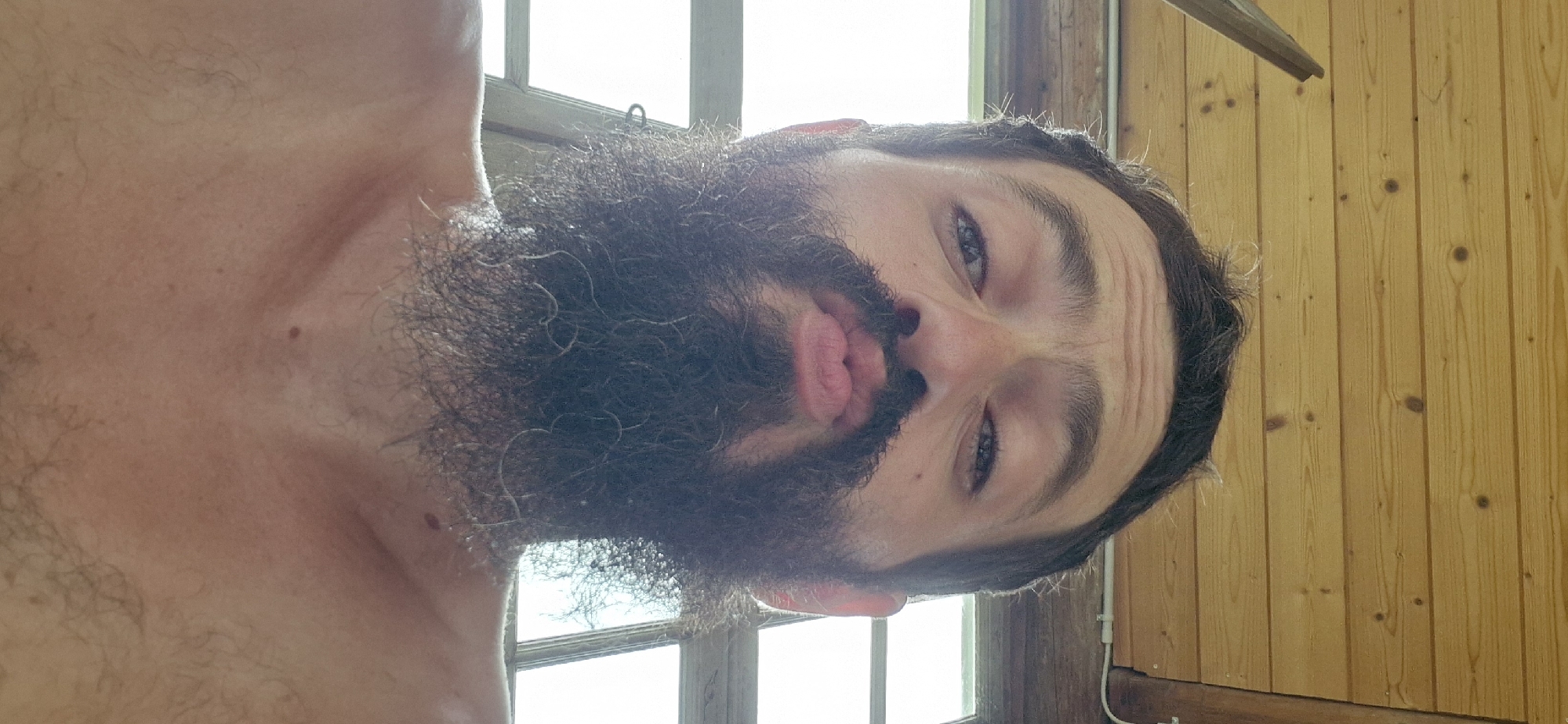 A close-up, low-angle selfie shows a man with a beard puckering his lips. The man's head and shoulders are visible, against a backdrop of wooden ceiling planks and a window. The image is taken in Wattenwil, Switzerland. The man is looking directly at the camera, with his eyes open and eyebrows furrowed. His beard is dark and bushy, with some lighter hairs scattered throughout. The man's skin is lightly tanned, and he has a few moles visible on his neck. The wooden planks are a light brown color, with visible knots and grain. The window is out of focus, but appears to be a large pane of glass.