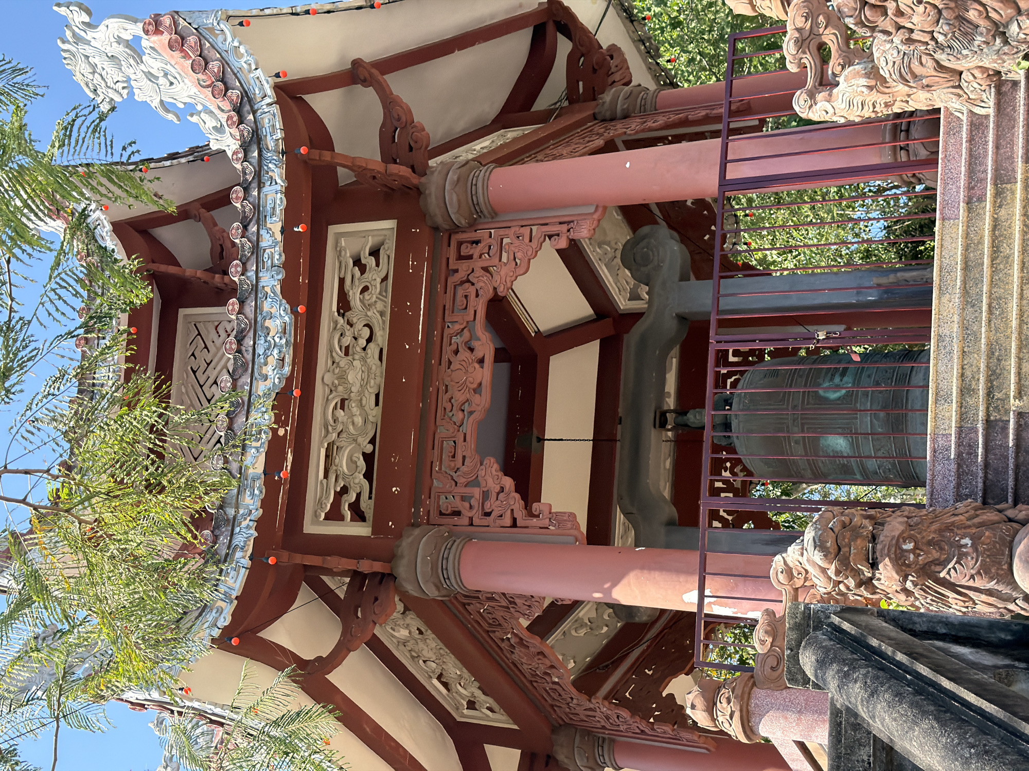 The image captures a section of a traditional Vietnamese architectural structure, likely a temple or shrine, in Tay Nha Trang Ward, Vietnam. The building features intricate carvings and embellishments in shades of red, white, and grey, possibly made of wood or stone. A large bronze bell hangs within the structure, enclosed by a protective iron cage. The architectural style is characterized by curved eaves, ornate roof decorations, and decorative support beams. Green foliage partially obscures the view of the building and the blue sky in the background.