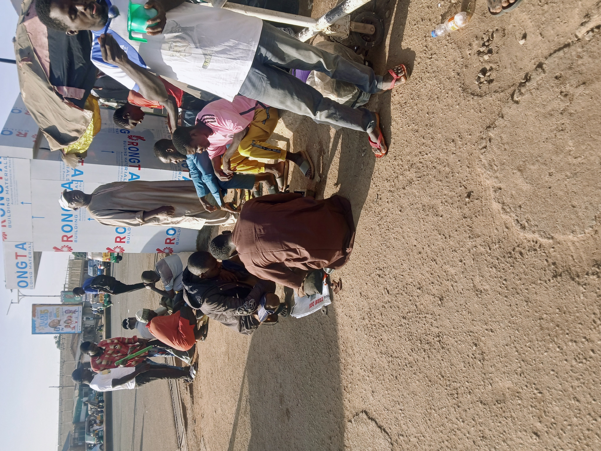 The image depicts a street scene in Gura Top, Nigeria, where a group of men are gathered. Some are seated on the ground, while others are standing. One man is wearing a light-colored robe and a white cap. A banner displays the words "Rongta Building Material" and "Ongta Aluminum Composite Panel." A poster advertising "Talent Unveiled 3" is visible in the background. The street is a sandy color.