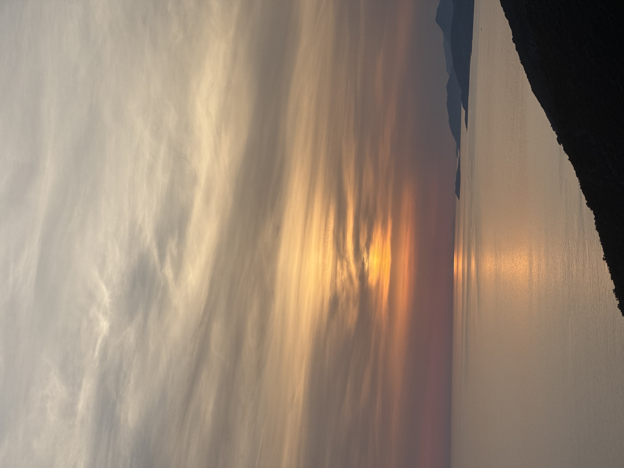 The image captures a serene sunset over the Adriatic Sea, viewed from a high vantage point on the coast near Zagora, Montenegro. The sky is a canvas of soft, muted colors, with wisps of clouds painted in shades of peach, rose, and pale gray. A sliver of the sun, a bright, glowing orb, is visible just above the horizon, casting a shimmering golden path across the tranquil water. In the distance, a chain of dark, silhouetted mountains forms a dramatic backdrop, their peaks softened by the twilight. The foreground is dominated by a steep, dark hillside, covered in the shadowy outlines of vegetation, suggesting a rugged, natural landscape. The overall impression is one of profound peace and natural beauty, a quintessential Mediterranean evening.