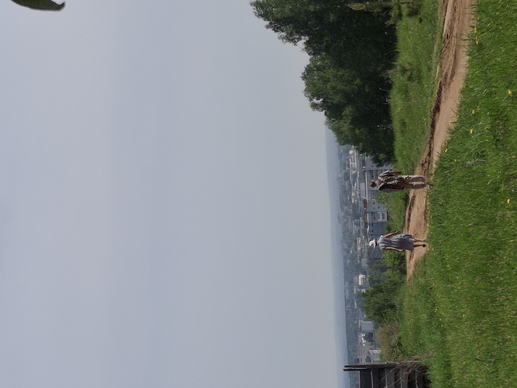 The image shows a view of the city of Chaikovsky, Russia from a high vantage point. A dirt path runs along the edge of a grassy slope. Two people are walking along the path. The sky is overcast and the city is partially obscured by haze. In the foreground, there is a section of a wooden fence.