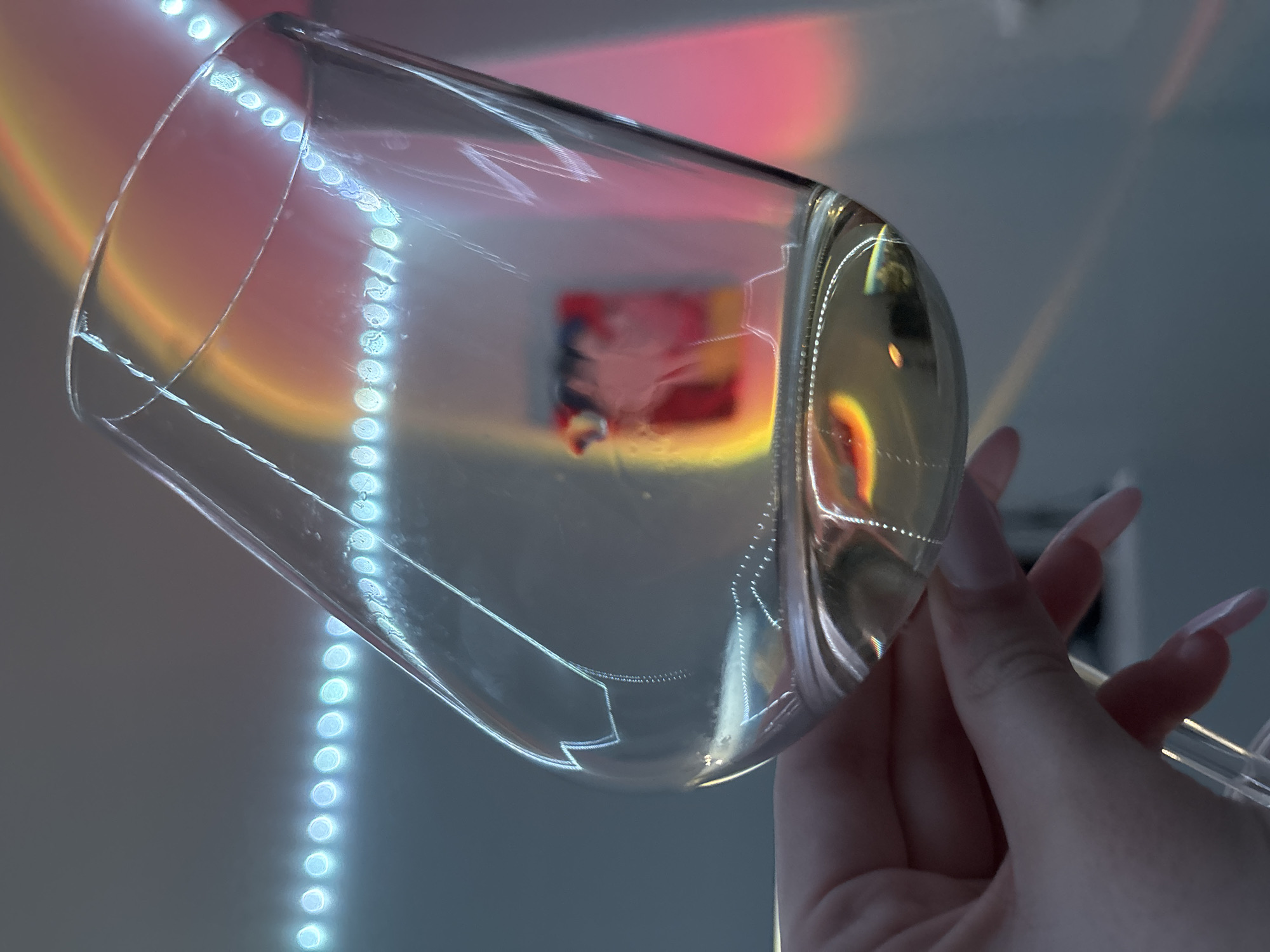 A close-up, low-angle shot depicts a clear wine glass held by a human hand. The hand, with pink artificial nails, grasps the stem of the glass. The glass is empty. A string of blue lights runs vertically in the background. Reflected through the glass is a colorful blurry image, possibly a picture or light source. The location is unknown. Russia is not indicated from the image.