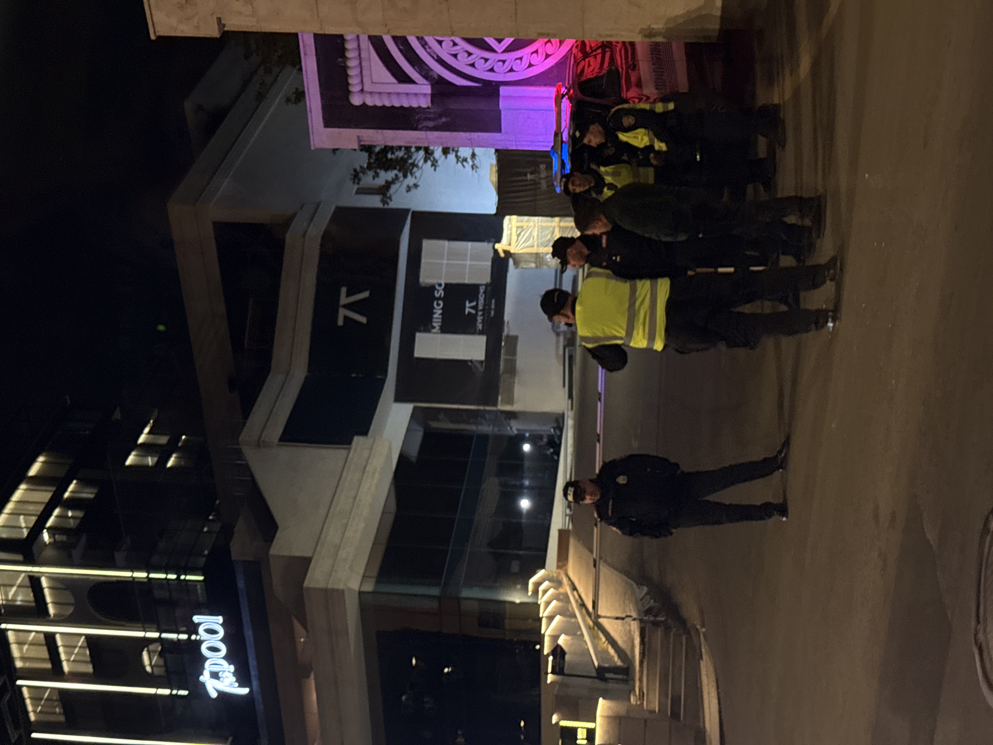 The photo shows a group of uniformed police officers lined up on a sidewalk at night in Yerevan, Armenia. One officer stands alone at the front of the line. The group is positioned in front of a modern building with prominent signage "7pool" and "COMING SOON". The architecture includes illuminated design elements. The street is lit, casting shadows from the officers and buildings. There are decorative architectural features above the group.