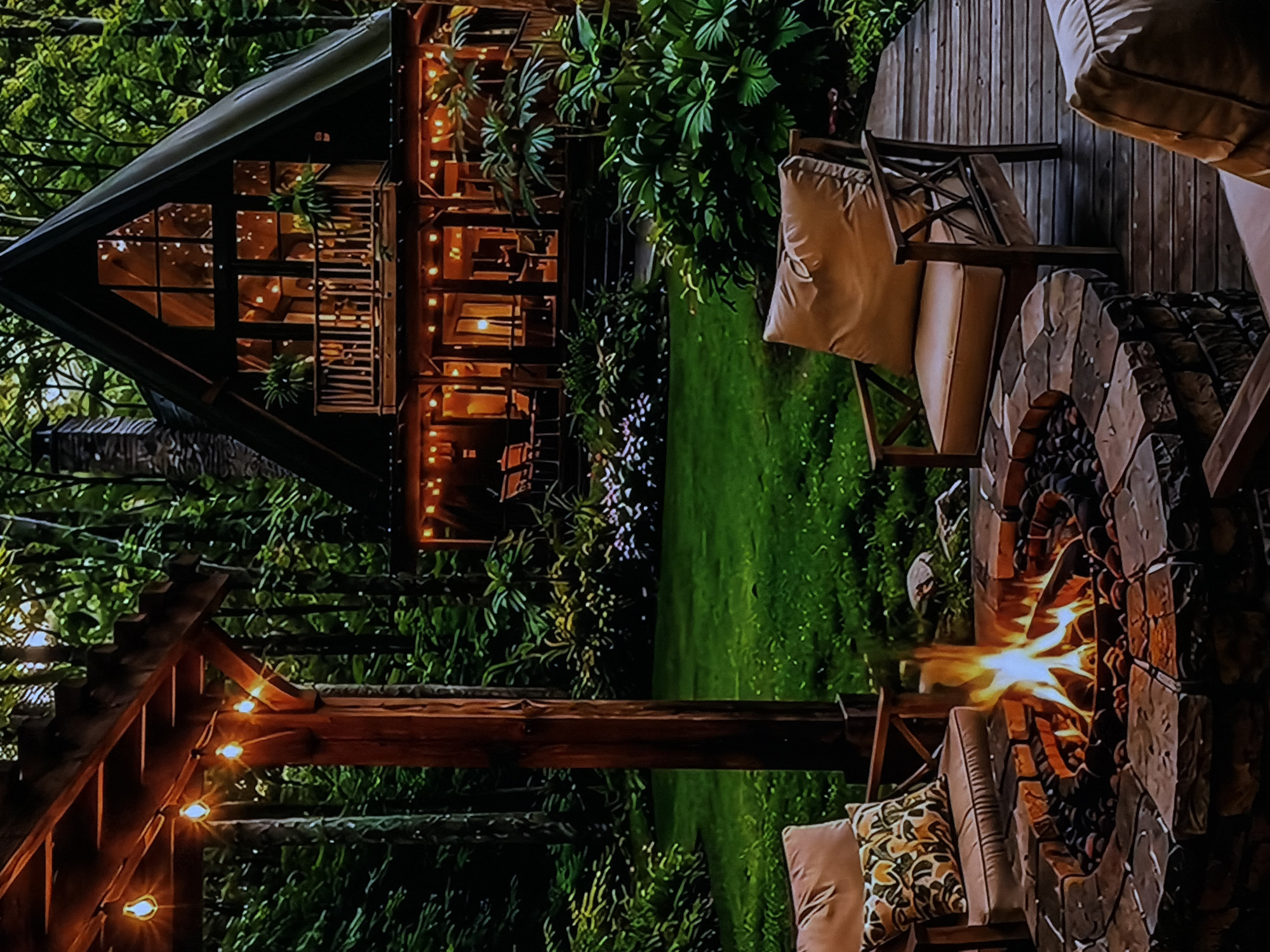 The image presents a serene nighttime scene in Sofiivska Borshchahivka, Ukraine, featuring a modern A-frame cabin and an inviting outdoor living area. The dark-sided cabin, possibly a retreat, stands against a backdrop of lush green trees, its numerous windows brightly illuminated, revealing interior activity and warmth. String lights decorate the cabin's balconies and exterior, enhancing its inviting glow.
In the foreground, a robust stone fire pit radiates warmth with active flames, serving as the central gathering point. Arranged around it on a wooden deck are comfortable outdoor chairs, furnished with thick, light brown cushions and patterned pillows, suggesting a space for relaxation and socializing. Overhead, a wooden pergola is adorned with string lights, casting a soft, ambient glow over the seating area. The entire setting is enveloped by dense green foliage and a grassy lawn, indicative of a peaceful, natural environment.
No individuals are present, but the arrangement strongly implies an atmosphere of coziness, tranquility, and an ideal setting for an evening gathering or quiet contemplation. The clear night sky is not visible, but the artificial illumination from the cabin, fire pit, and string lights confirms the time of day. There is no visible text in the image.