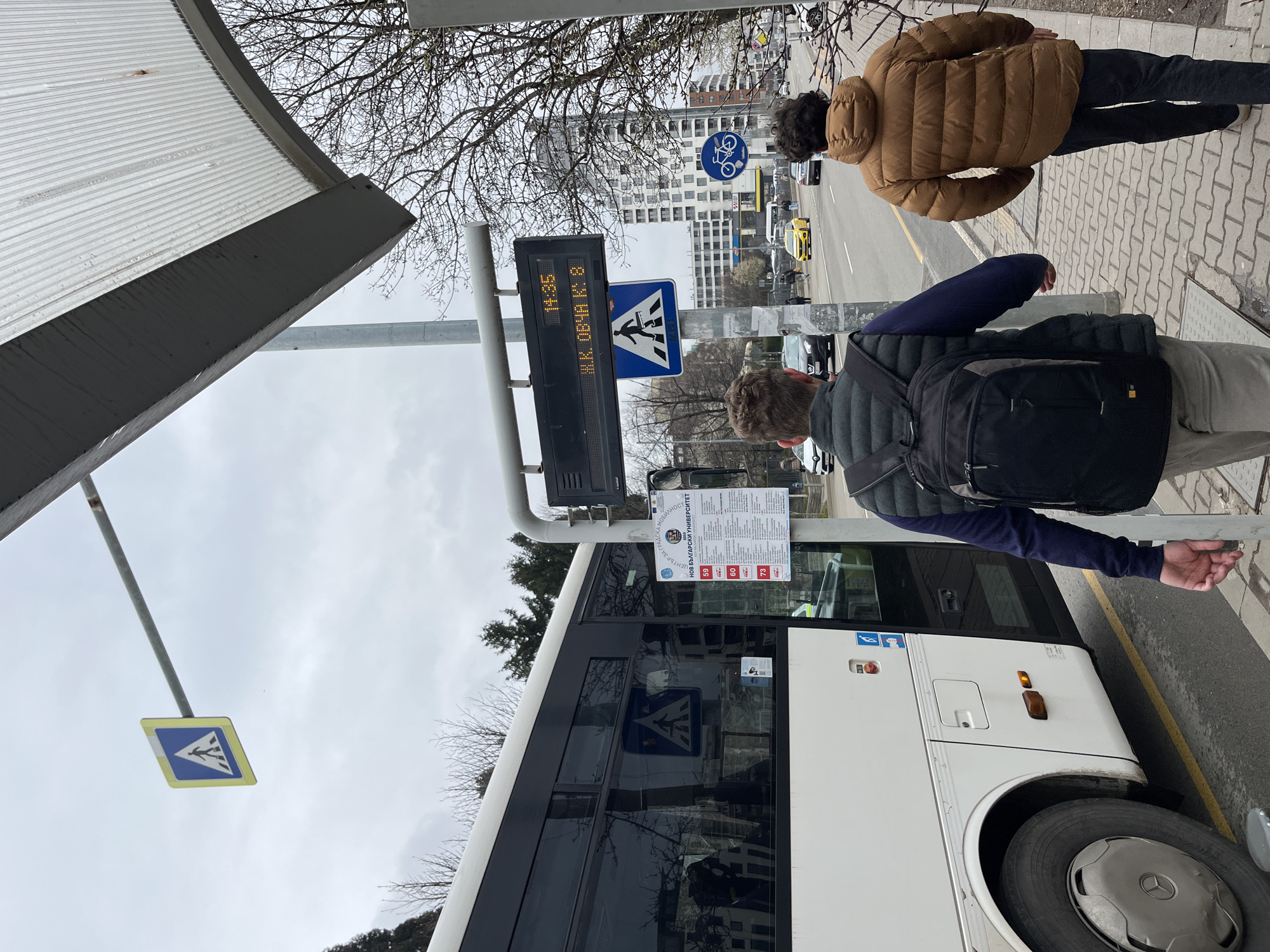 The image shows a bus stop in the city of Sofia, Bulgaria, likely near the New Bulgarian University (НБУ) as indicated by the text on the bus stop sign "НОВ БЪЛГАРСКИ УНИВЕРСИТЕТ". A white bus is parked at the stop. Two people are standing on the sidewalk waiting. One person wears a brown jacket and black pants, and the other person wears a gray pants, a dark blue sweater and a dark green vest, and a black backpack. The sign at the bus stop displays the time as 14:35 and the route is Ж.К. ОВЧА К: 8. A pedestrian crossing sign is also visible. A street and buildings are visible in the background.