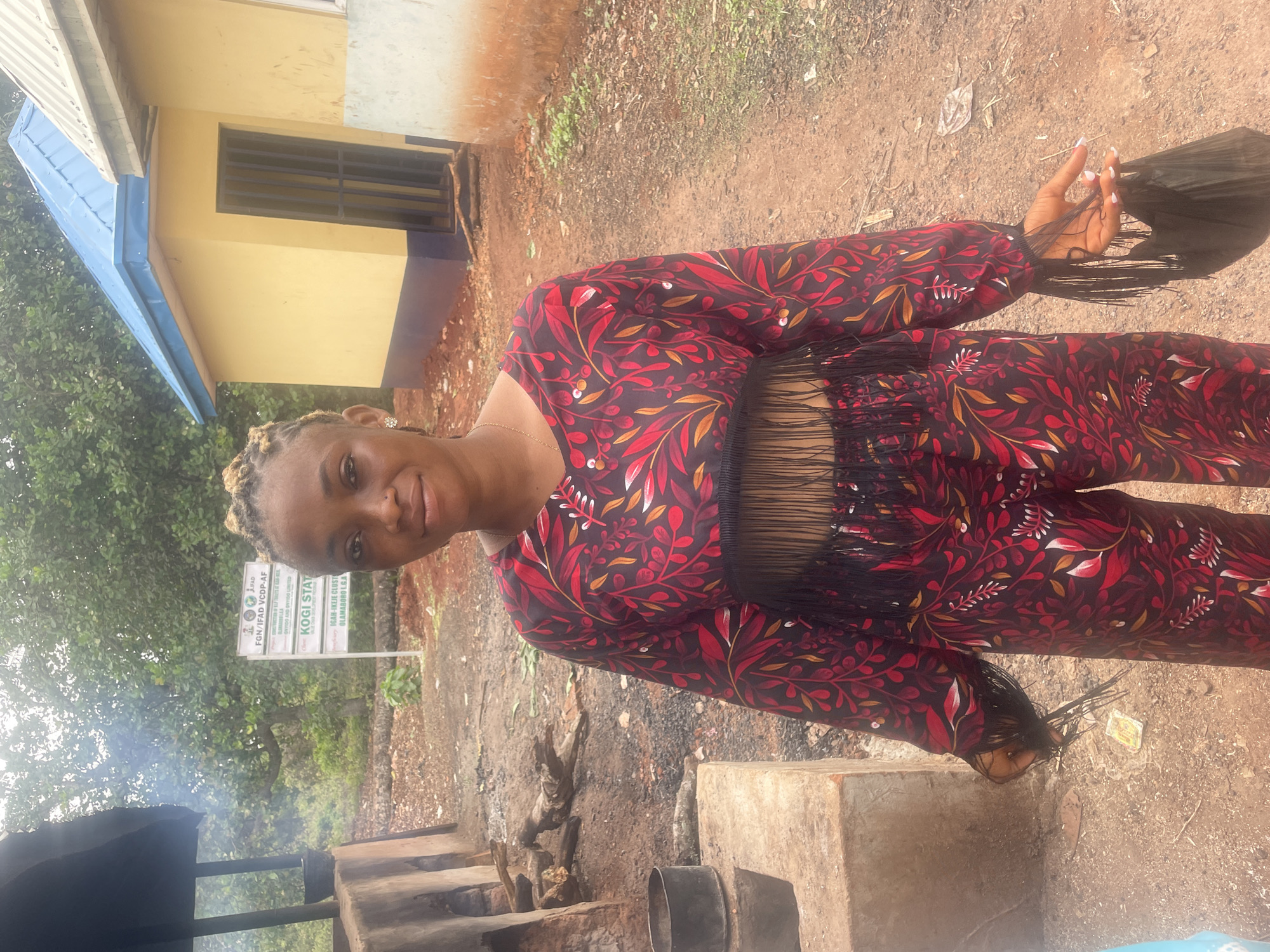 A woman stands outside in Eke, Nigeria. She wears a red and black patterned outfit with fringed detailing. Behind her is a yellow building with a blue roof and barred windows. To her left, smoke rises near what appears to be a cooking area constructed from cinder blocks and metal containers. A sign is visible in the background with the text "FGN/IFAD VCDP-AF," "ONTIDO AND ONTIDO LIMITED," and "KOGI STAT." The ground is a mix of dirt and vegetation.