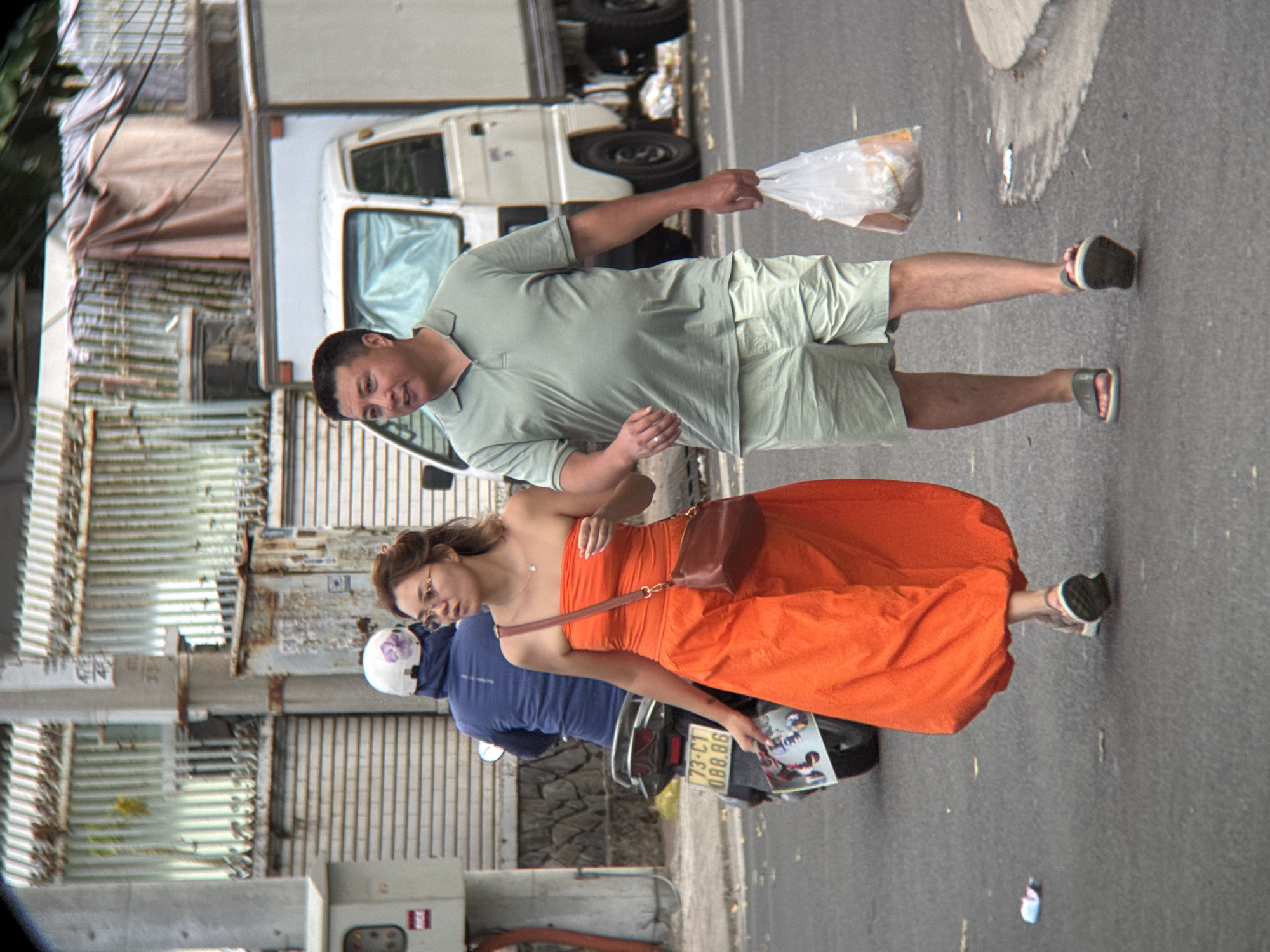 An outdoor, eye-level, medium shot captures two adults walking on a paved road in Nha Trang, Vietnam. The woman on the left, wearing a bright orange strapless dress and sunglasses, walks with her right hand slightly raised, holding a brown purse on her left shoulder. She appears to be looking to her right. To her right, a man in a pale green polo shirt and light-colored shorts walks with a plastic bag containing food in his right hand. He is looking forward. Behind them, a person on a motorcycle, wearing a blue jacket and a white helmet with purple floral patterns, is visible. The background features a corrugated metal fence, a white truck, and some debris on the road. The weather appears overcast.