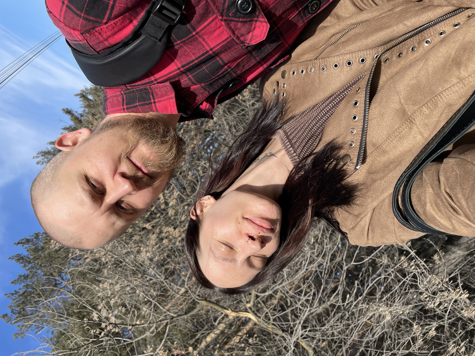 A photo shows two individuals outdoors. The person on the left has a shaved head and a beard. They are wearing a red and black plaid shirt with a black shoulder bag strap across their chest. The person on the right has long, dark hair. They are wearing a brown jacket with metal eyelets. Behind them is a background of bare branches and a blue sky. There is no definitive information in the image to determine the location, specifically within the city of Izhevsk, Russia.