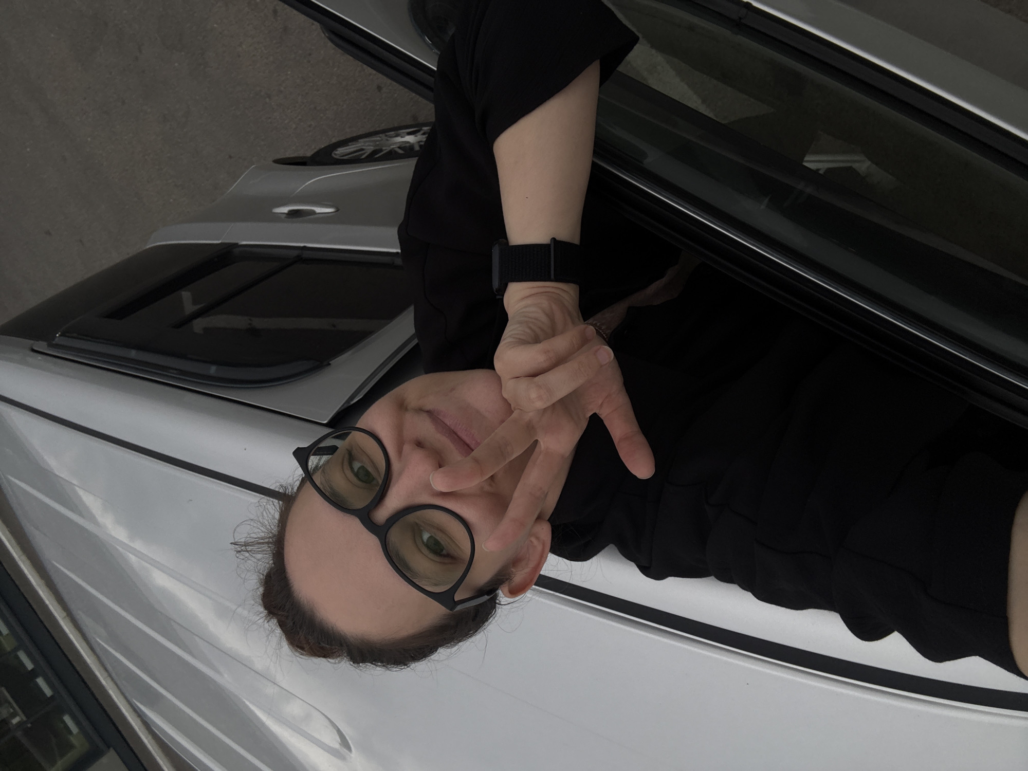 A person with glasses is seated inside a silver car, partially visible through the open car door. The person is making a "peace" or "victory" sign with their fingers. They are wearing a black shirt and a watch. The car has a sunroof. The location is unknown.