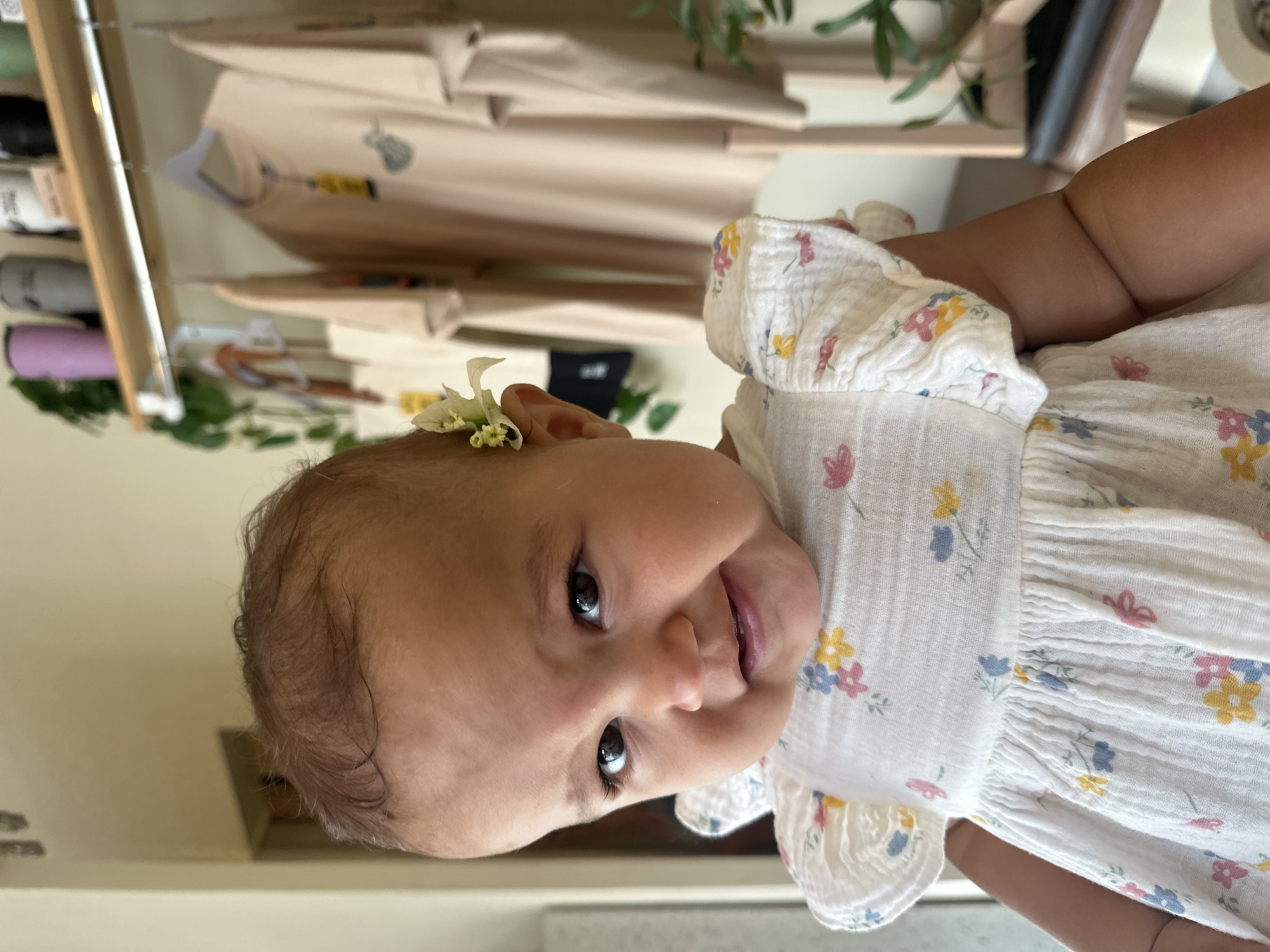 A close-up of a baby, perhaps 6-12 months old, with light brown skin and dark brown eyes. The baby has thin, dark brown hair. A small, white flower is tucked behind the baby's left ear. The baby wears a white dress with a floral print featuring small blue, yellow, and pink flowers. The baby's expression is a slight smile. In the blurred background, there are clothes hanging on a rack, greenery, and other items. The location appears to be indoors. There is no information in the image to suggest that it was taken in North Kuta, Indonesia.