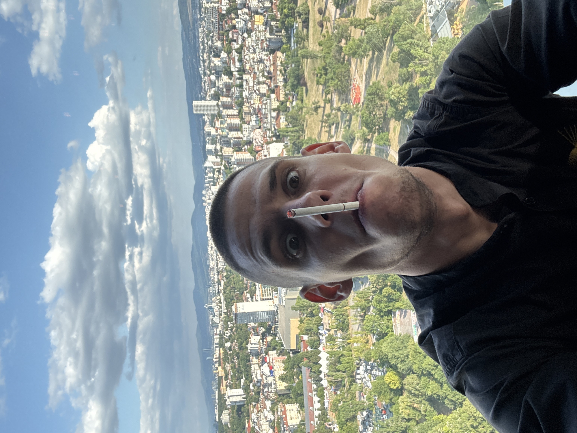 A man is taking a selfie from a high vantage point overlooking a city. He has a lit cigarette balanced vertically on his nose. He is wearing a black collared shirt and his expression is one of exaggerated surprise. The background shows a dense urban landscape with numerous buildings of varying heights and architectural styles, interspersed with green trees. In the distance, mountains are visible under a partly cloudy sky. The city appears to be Sofia, Bulgaria, based on the presence of the National Palace of Culture (NDK) and its surrounding area, which is a prominent landmark. The foreground is dominated by the man's face, with the city and sky forming the backdrop.